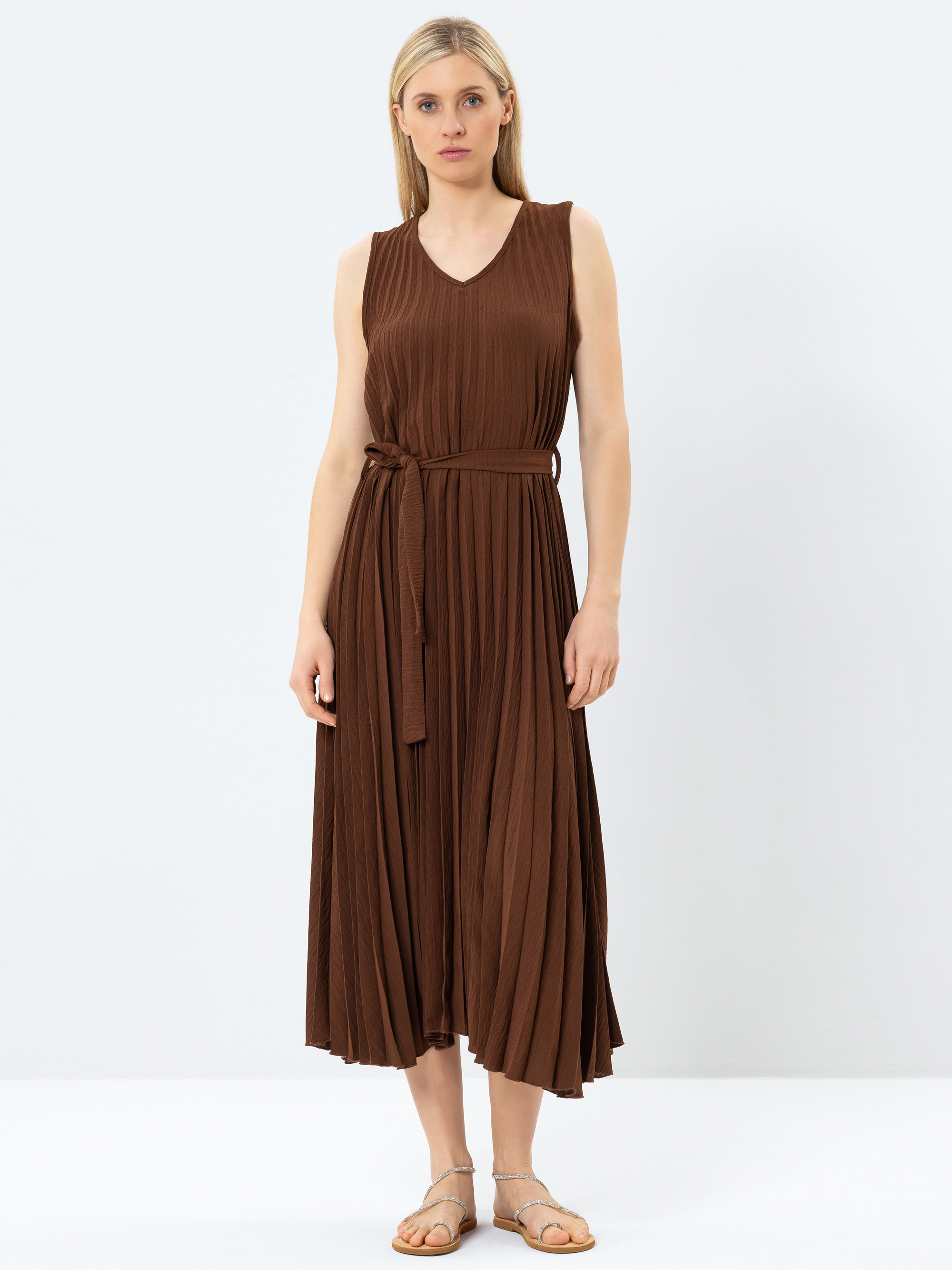 Eine Frau steht vor einem schlichten weißen Hintergrund und trägt ein ärmelloses, braunes, plissiertes Midikleid mit einem gebundenen Taillengürtel und flachen Sandalen. Sie hat glattes blondes Haar und einen neutralen Gesichtsausdruck.