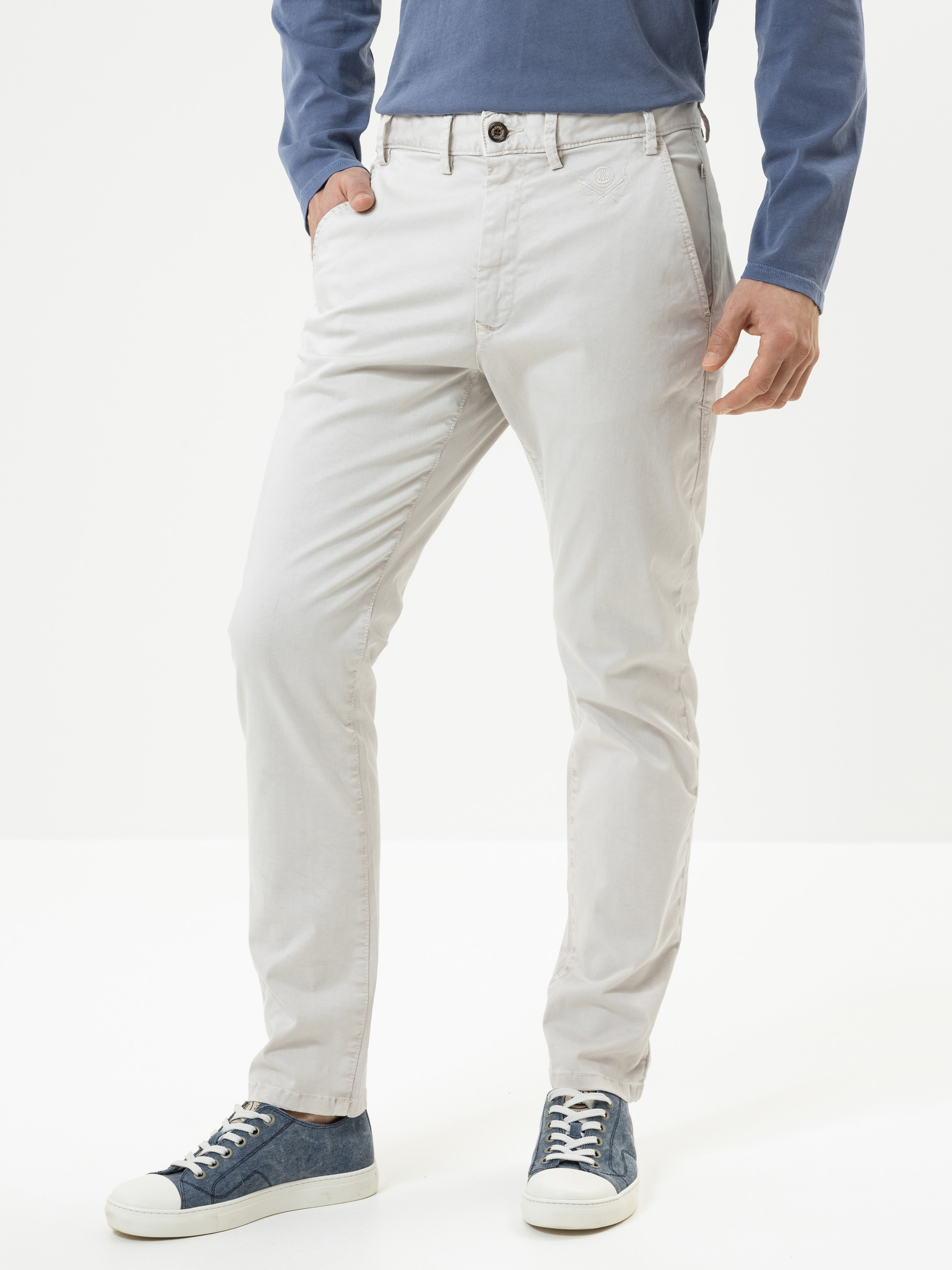 Eine Person, die eine hellgraue Chinohose, ein blaues langärmeliges Hemd und blau-weiße Turnschuhe trägt, steht mit einer Hand in der Tasche vor einem schlichten weißen Hintergrund.