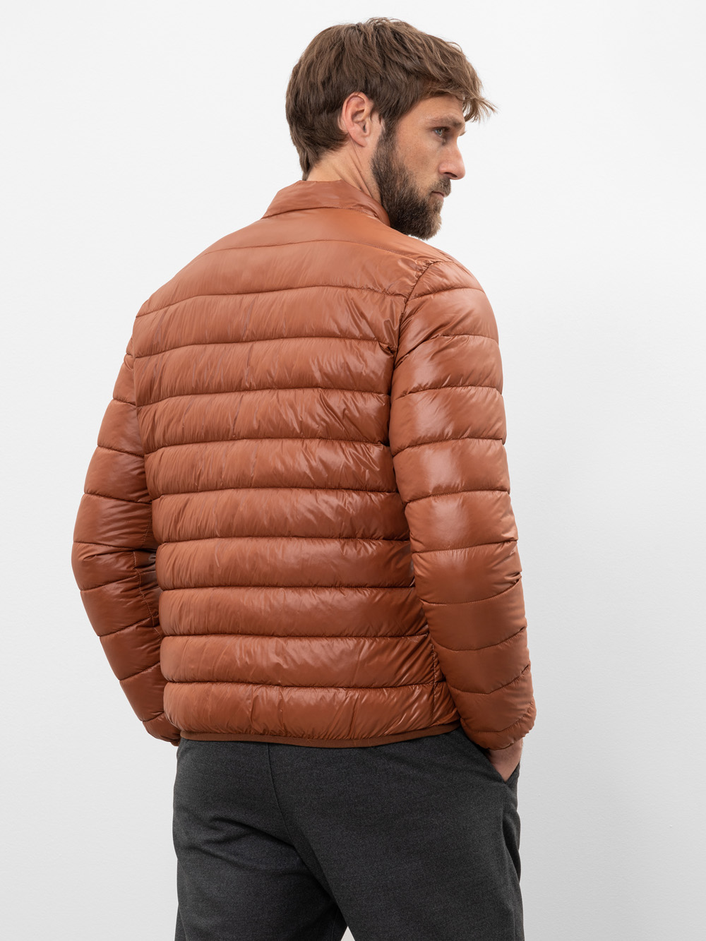 Ein Mann mit kurzen braunen Haaren und einem Bart trägt eine braune Pufferjacke und eine dunkle Hose und steht mit dem Rücken zur Kamera vor einem schlichten weißen Hintergrund.