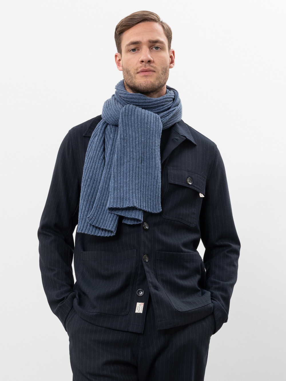 Ein Mann in einer dunklen Nadelstreifenjacke und -hose steht mit den Händen in den Taschen. Er hat einen ernsten Gesichtsausdruck und trägt einen dicken, gerippten blauen Schal um den Hals gewickelt. Der Hintergrund ist einfarbig weiß.