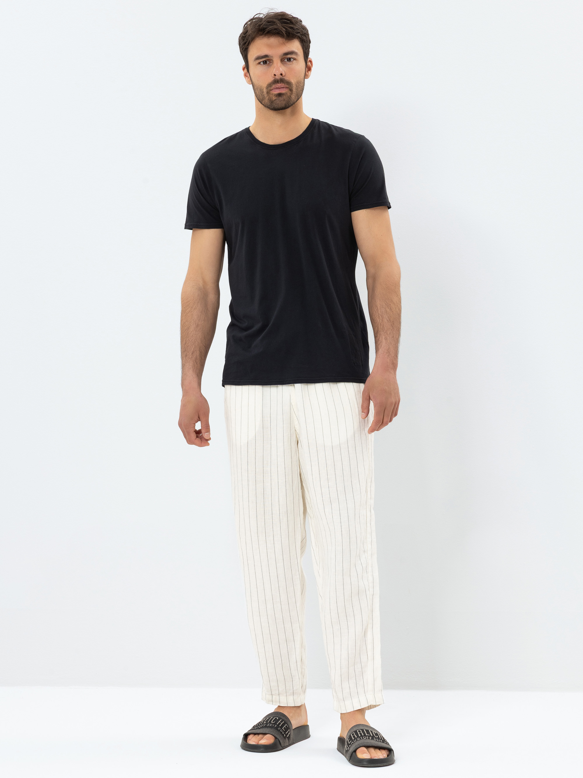 Ein Mann steht vor einem schlichten weißen Hintergrund und trägt ein schwarzes T-Shirt, eine cremefarbene Hose mit dünnen vertikalen Streifen und schwarze Slide-Sandalen.