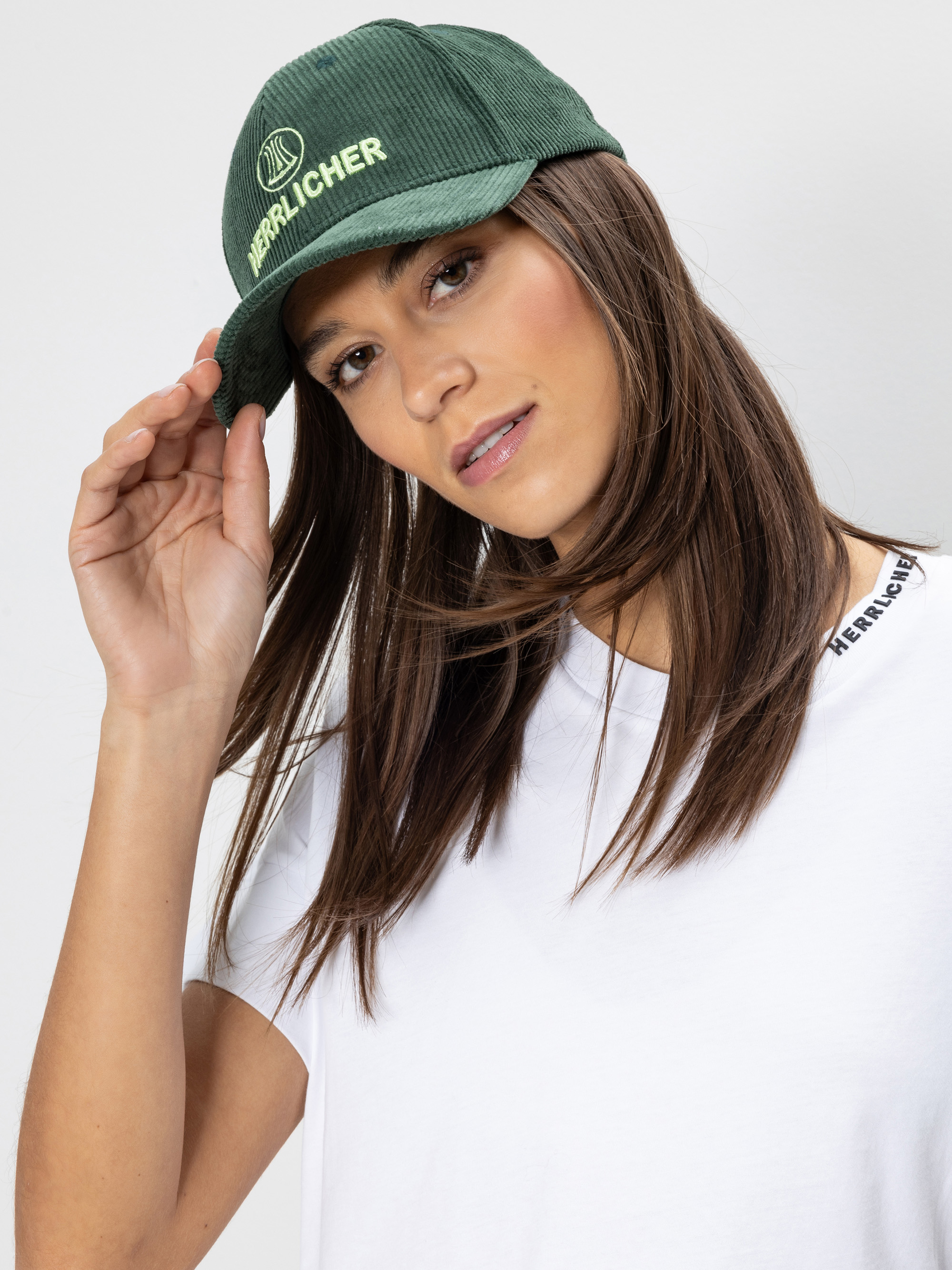 Eine Frau mit glattem braunem Haar trägt ein weißes T-Shirt und eine grüne Mütze mit der Aufschrift "HERZLICHER". Sie neigt den Kopf leicht und hält die Krempe ihrer Mütze, während sie in die Kamera schaut.
