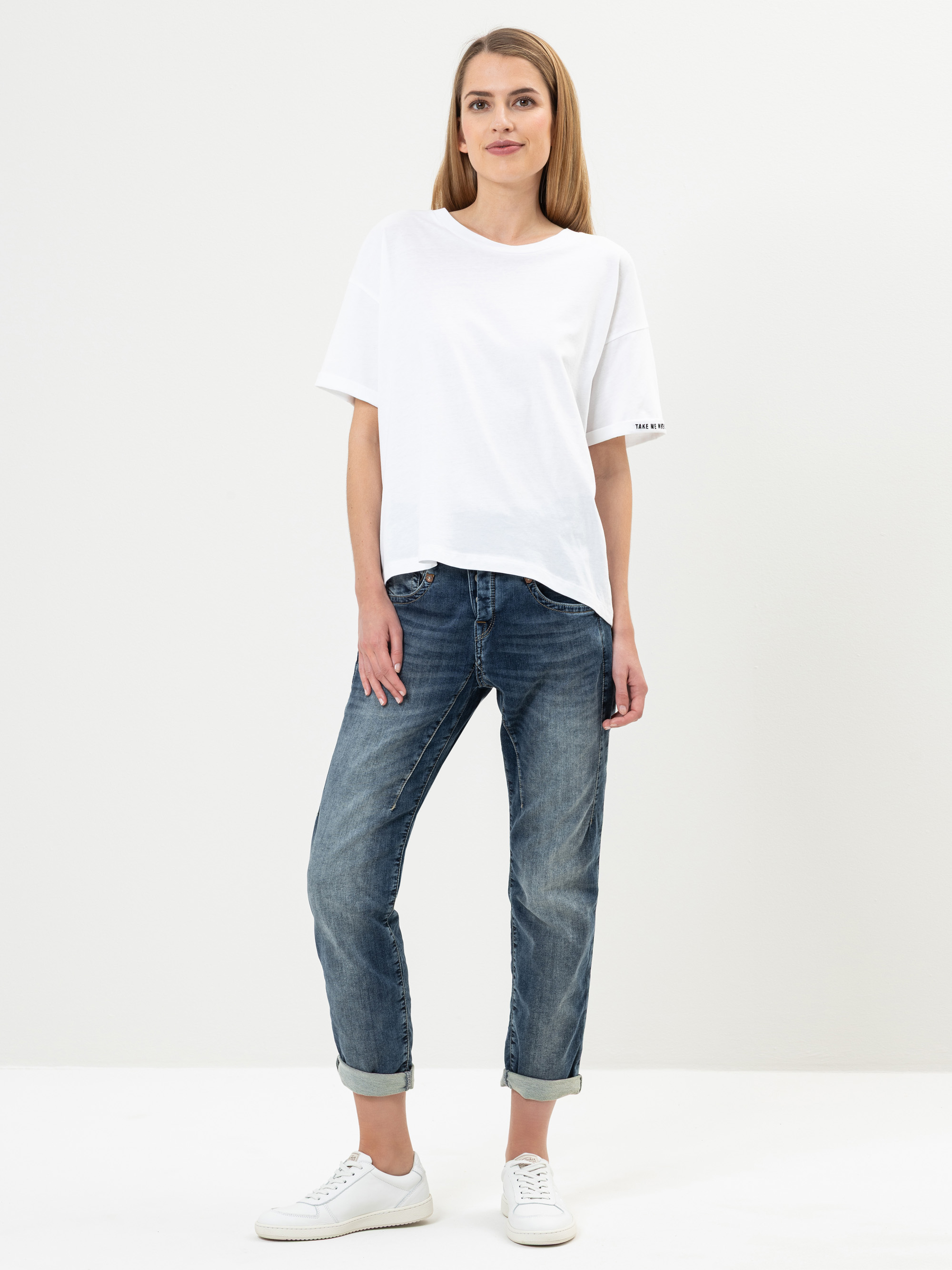 Eine Frau steht vor einem schlichten weißen Hintergrund und trägt ein lockeres weißes T-Shirt, blaue Jeans mit Bündchen und weiße Turnschuhe. Sie hat glattes, hellbraunes Haar und einen neutralen Gesichtsausdruck.