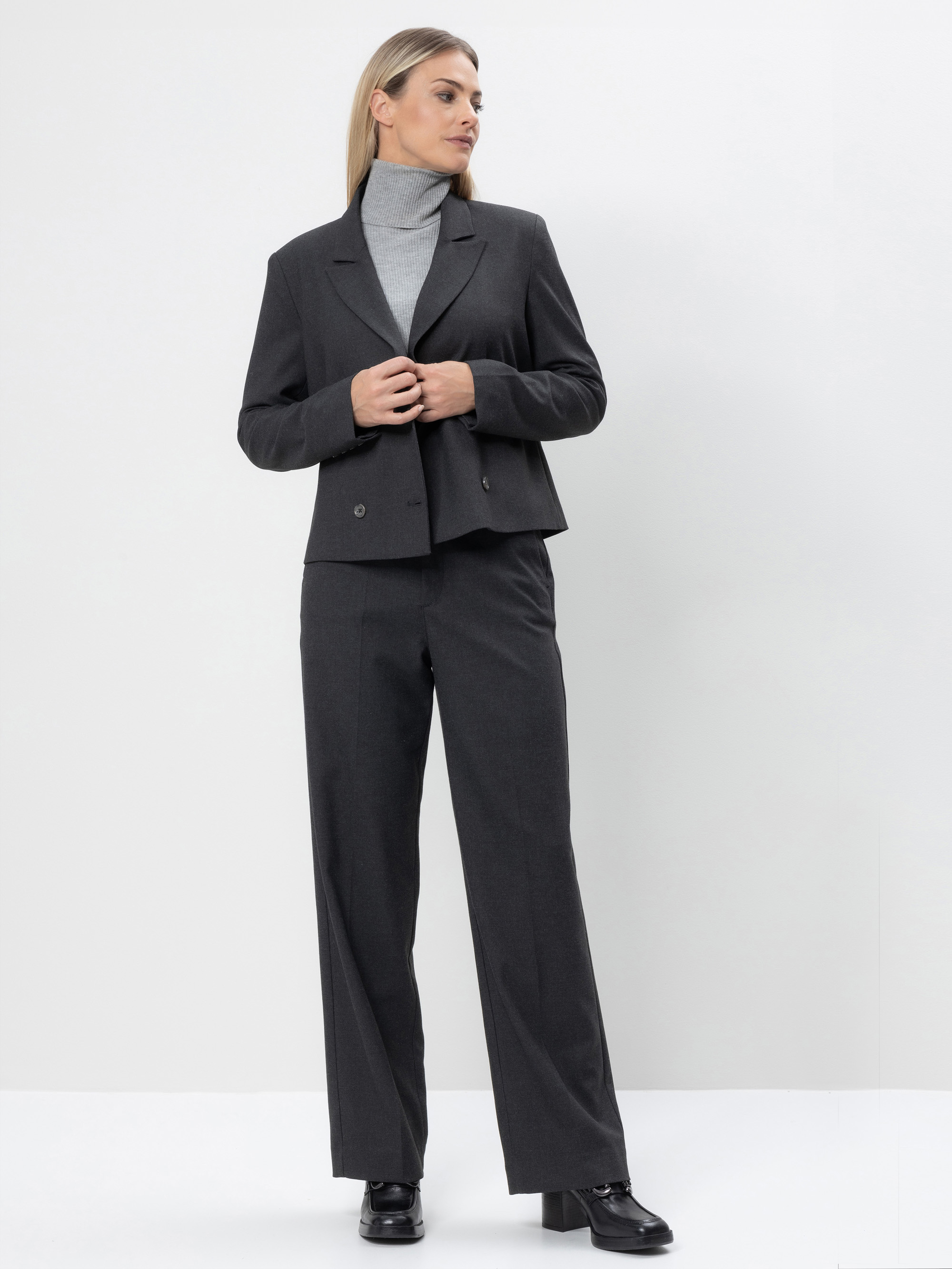 Eine Frau steht vor einem schlichten weißen Hintergrund, trägt einen dunkelgrauen Hosenanzug mit einem grauen Rollkragenpullover und schwarzen Schuhen und schaut zur Seite, während sie ihren Blazer in den Händen hält.
