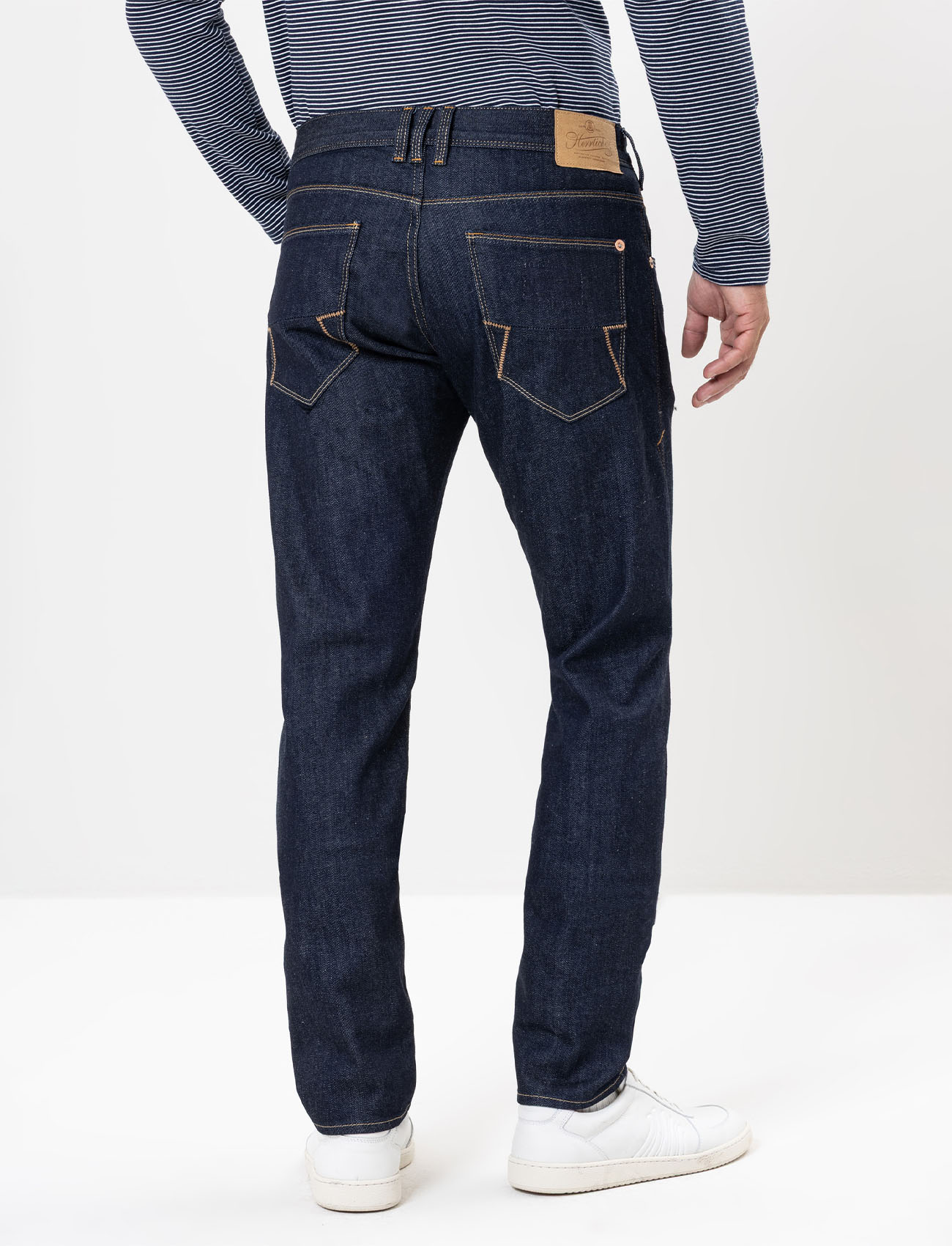 Eine Person, die dunkelblaue Jeans, ein blau-weiß gestreiftes langärmeliges Hemd und weiße Turnschuhe trägt, wird von hinten vor einem schlichten hellen Hintergrund gezeigt.