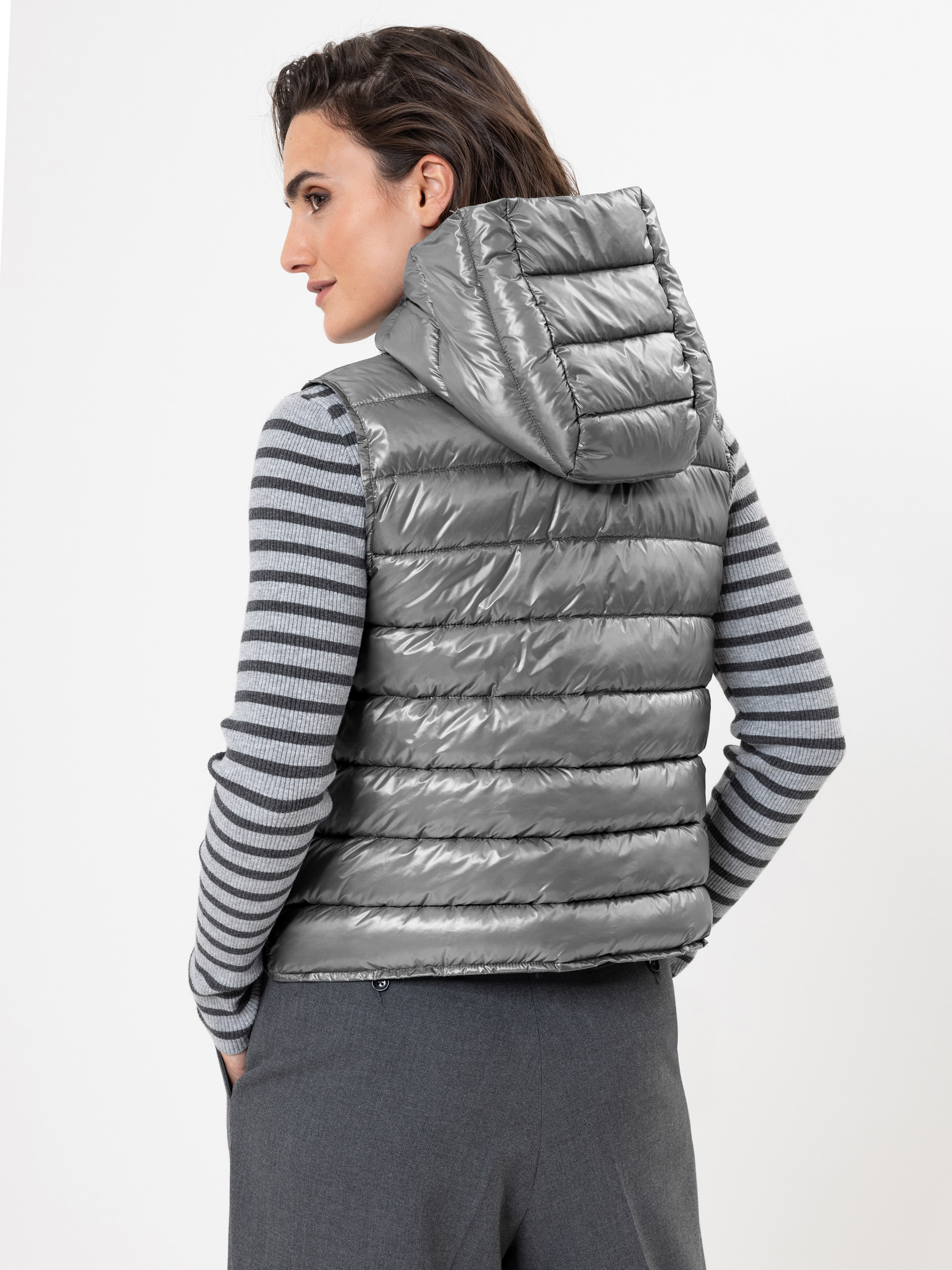 Eine Person, die eine silbern glänzende Kapuzenweste über einem grau-weiß gestreiften Pullover trägt und leicht von der Kamera wegschaut, vor einem schlichten weißen Hintergrund.