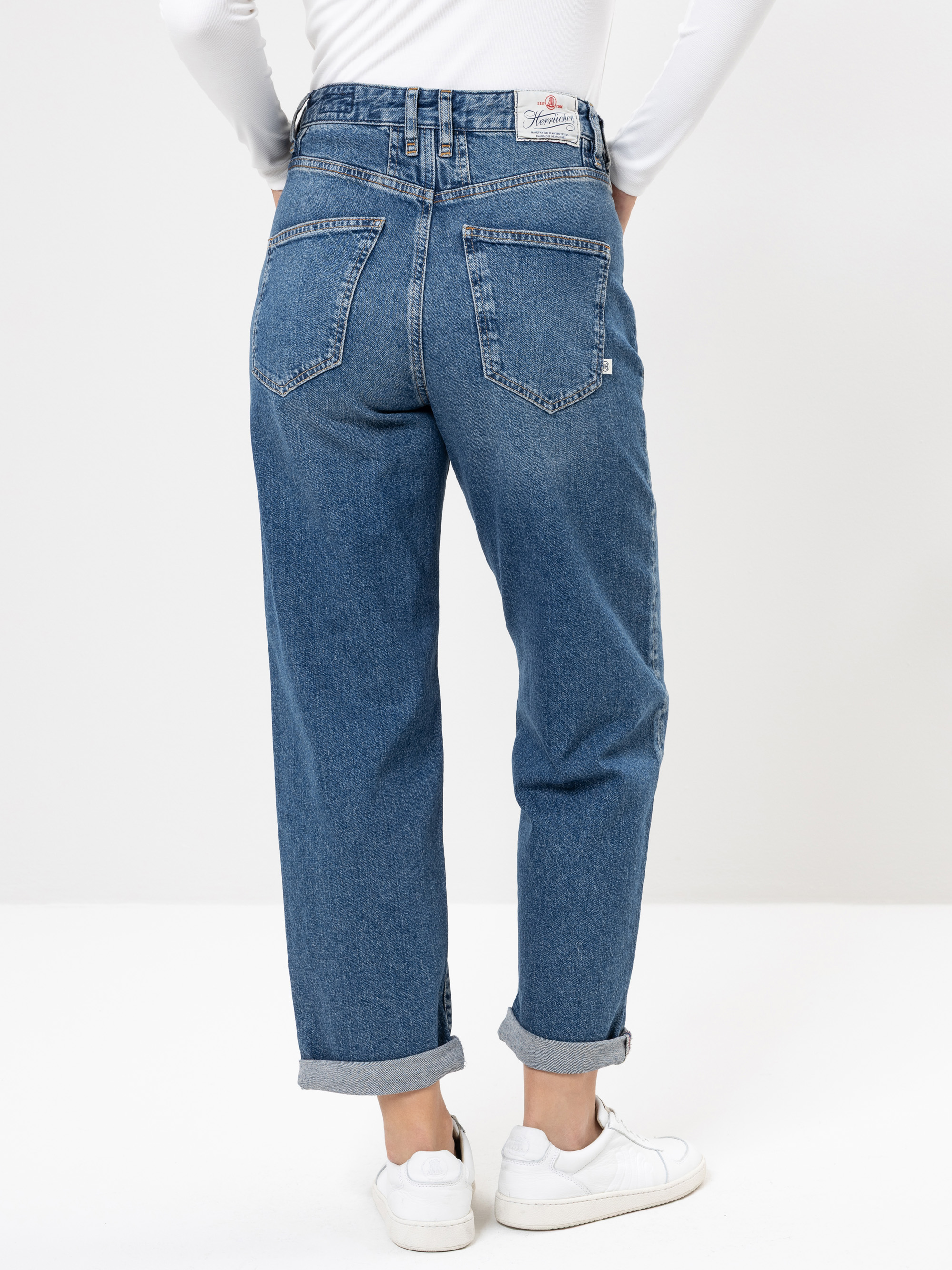 Eine Person, die hochgekrempelte blaue Jeans, ein weißes langärmeliges Oberteil und weiße Turnschuhe trägt, von hinten vor einem schlichten hellen Hintergrund.