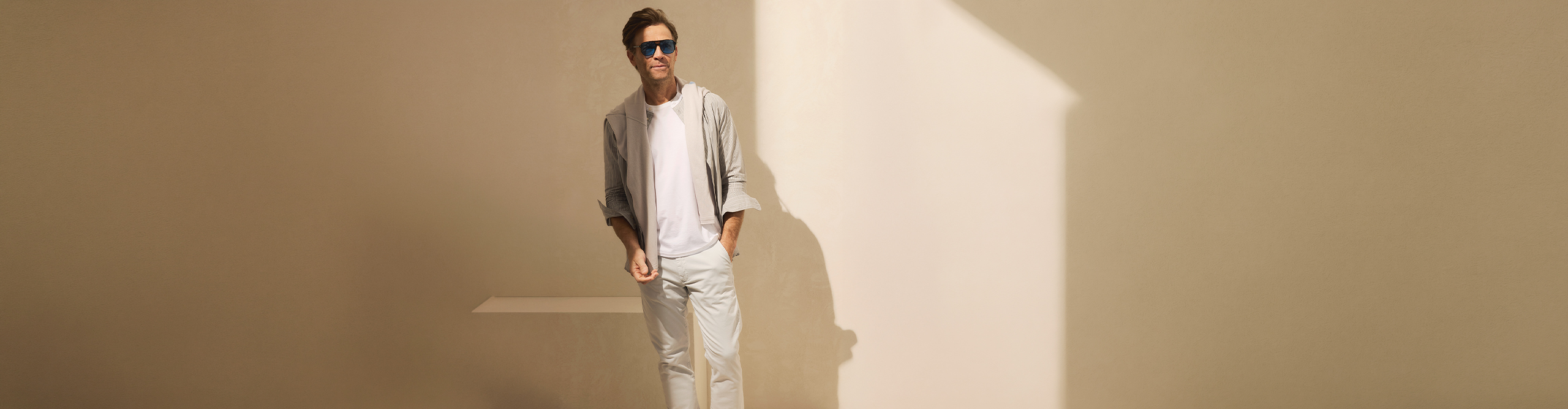 Ein Mann, der eine Sonnenbrille, ein weißes T-Shirt, eine helle Hose und eine helle Jacke trägt, steht vor einer beigen Wand, teilweise im Sonnenlicht, und hat eine Hand in der Tasche.