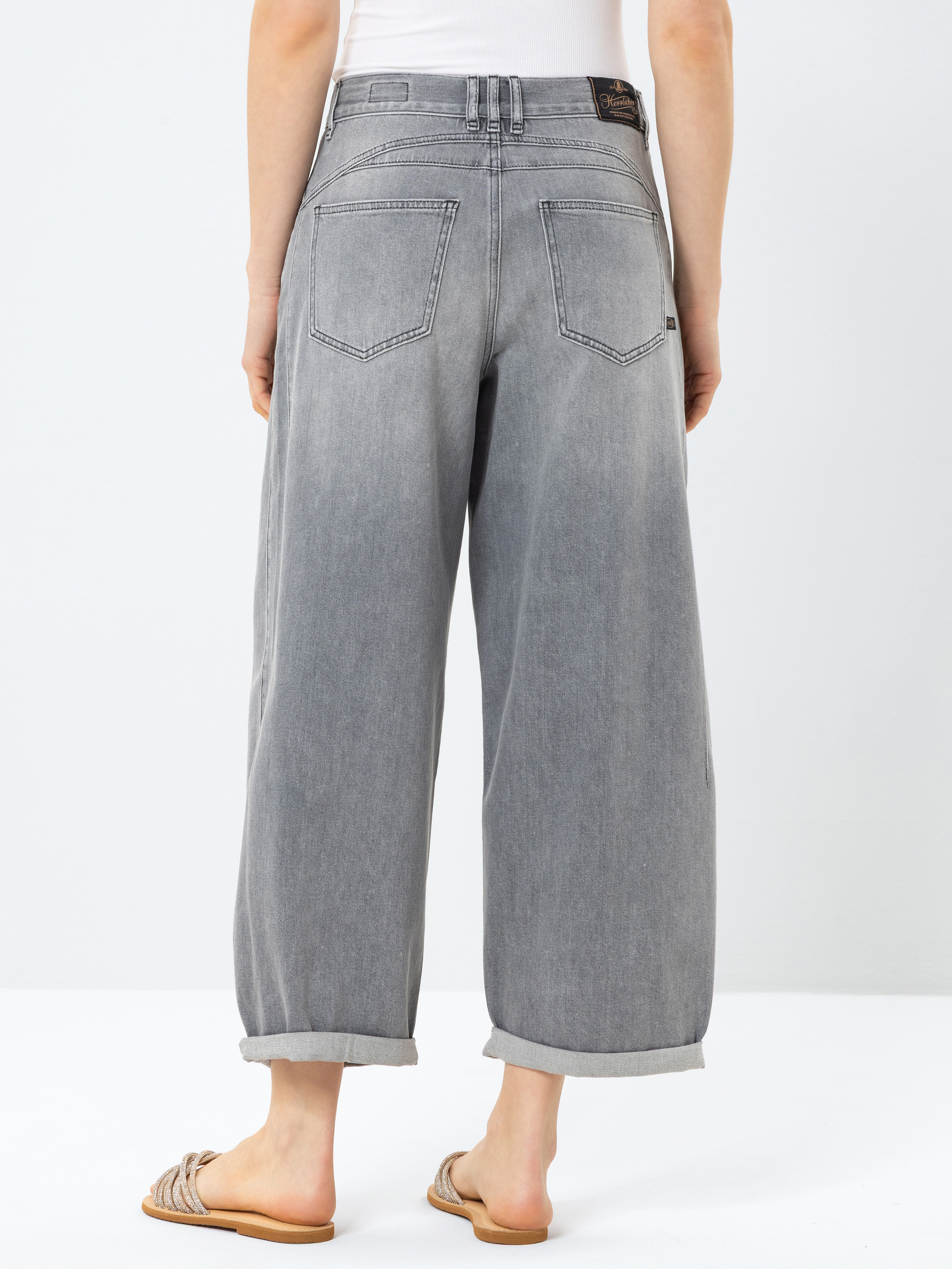 Eine Person, die eine hellgraue Jeans mit weitem Bein und hochgekrempelten Manschetten sowie ein weißes Oberteil trägt, steht mit dem Gesicht zur Seite. Sie trägt außerdem beigefarbene Sandalen auf weißem Grund.