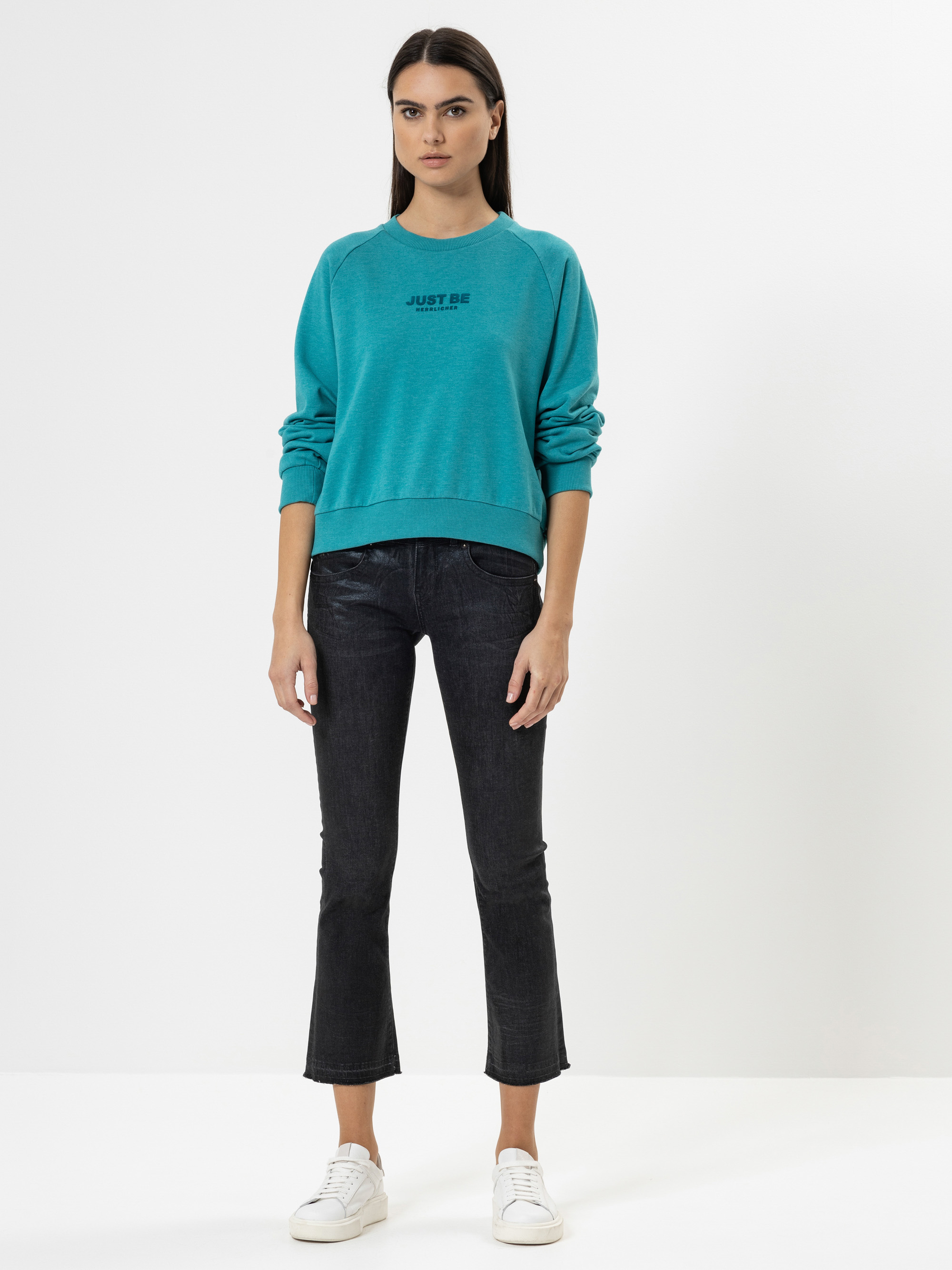 Eine Frau steht mit dem Gesicht nach vorne vor einem schlichten weißen Hintergrund und trägt ein blaugrünes Sweatshirt mit der Aufschrift JUST BE, dunkle Jeans und weiße Turnschuhe. Sie hat glattes dunkles Haar und einen neutralen Gesichtsausdruck.