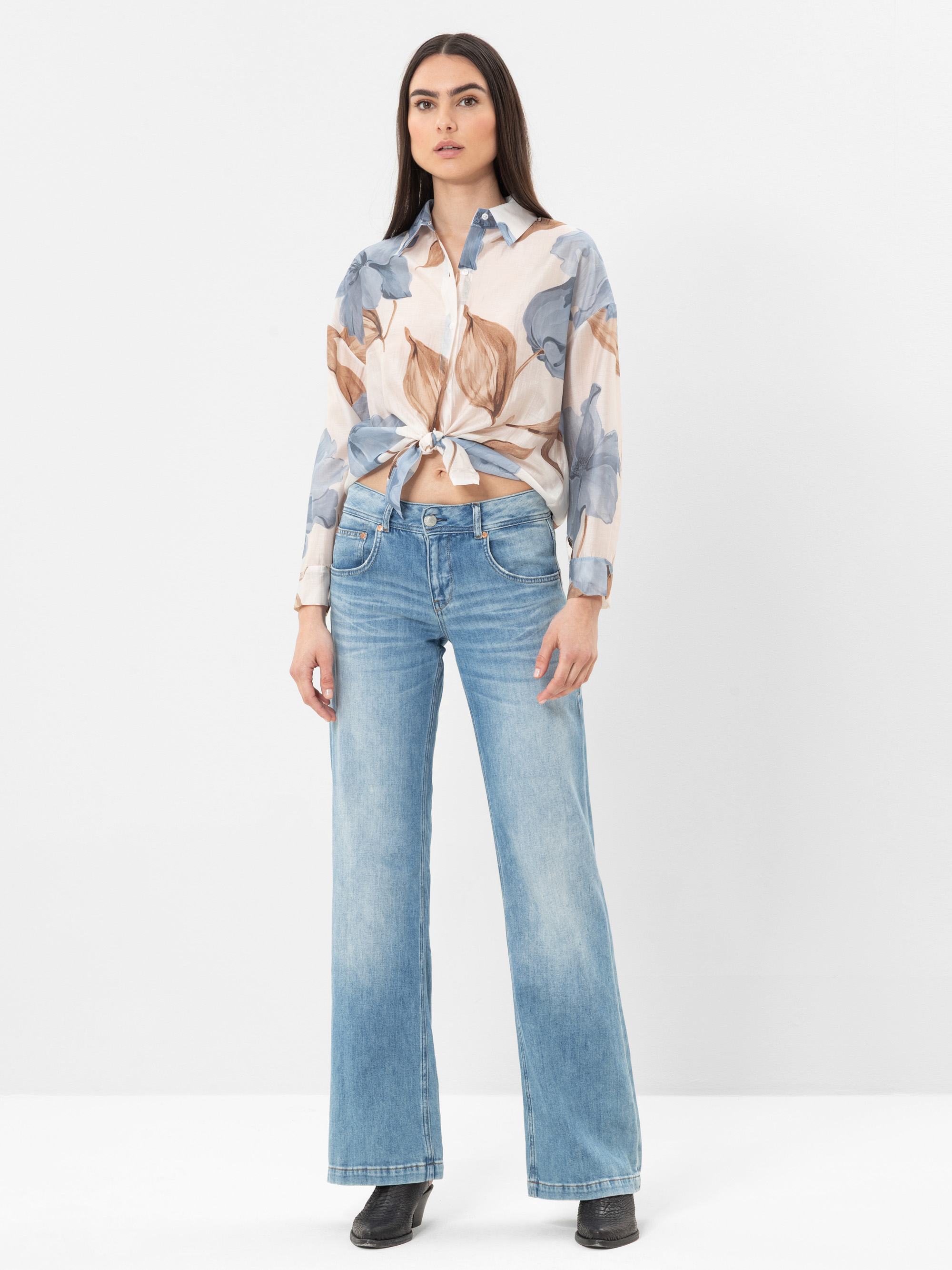Eine Frau steht vor einem weißen Hintergrund und trägt eine blaue Jeans mit weitem Bein, ein langärmeliges geblümtes Hemd, das in der Taille gebunden wird, und schwarze Schuhe. Sie hat langes dunkles Haar und einen neutralen Gesichtsausdruck.