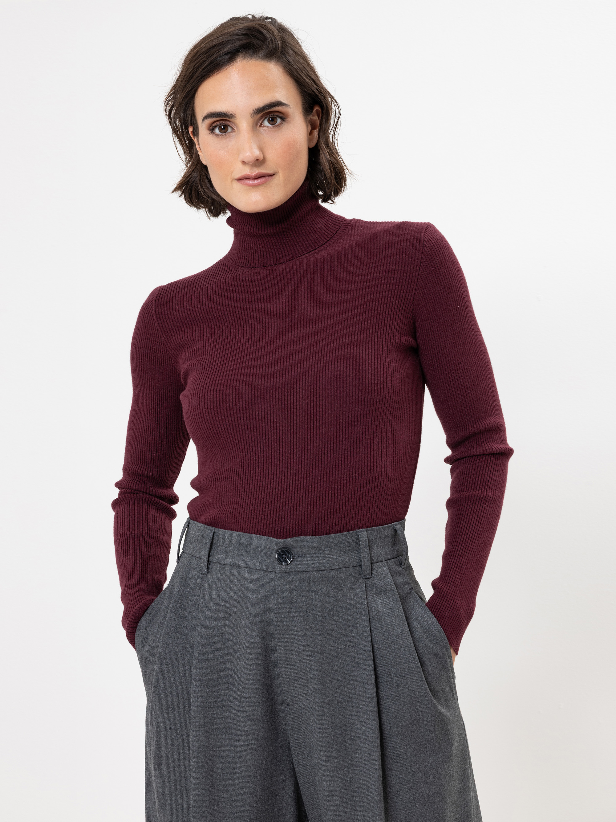Eine Person mit kurzen dunklen Haaren, die einen bordeauxroten Rollkragenpullover und eine graue Hose mit hoher Taille trägt, steht vor einem schlichten weißen Hintergrund, hat die Hände in den Taschen und einen neutralen Gesichtsausdruck.