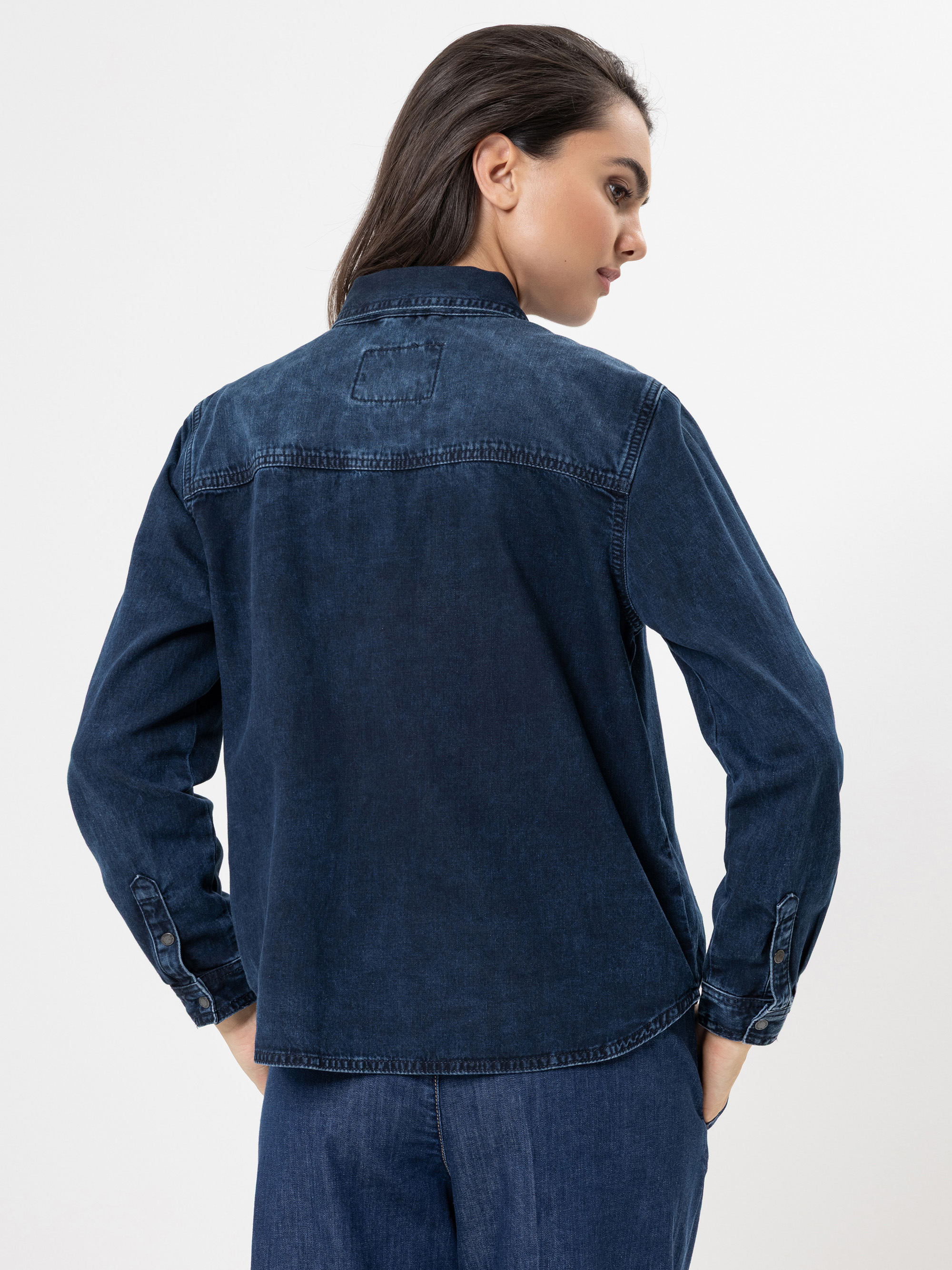 Eine Frau mit langen dunklen Haaren trägt ein dunkelblaues Jeanshemd und dazu passende Jeans. Sie steht mit dem Rücken zur Kamera und blickt über ihre Schulter vor einem schlichten weißen Hintergrund.