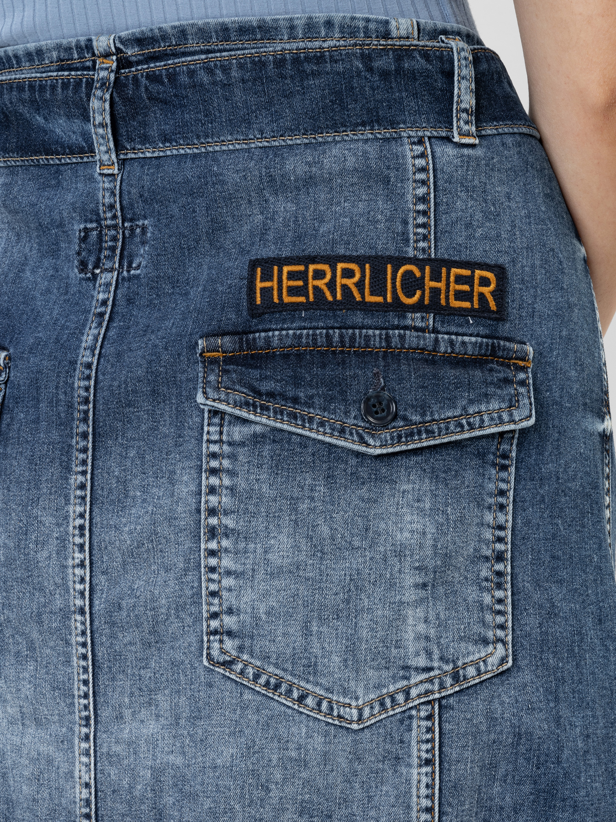 Nahaufnahme des Rückenteils eines blauen Jeansrocks mit einer Pattentasche. Über der Tasche befindet sich ein schwarz-gelber Aufnäher, auf den das Wort HERRLICHER gestickt ist.