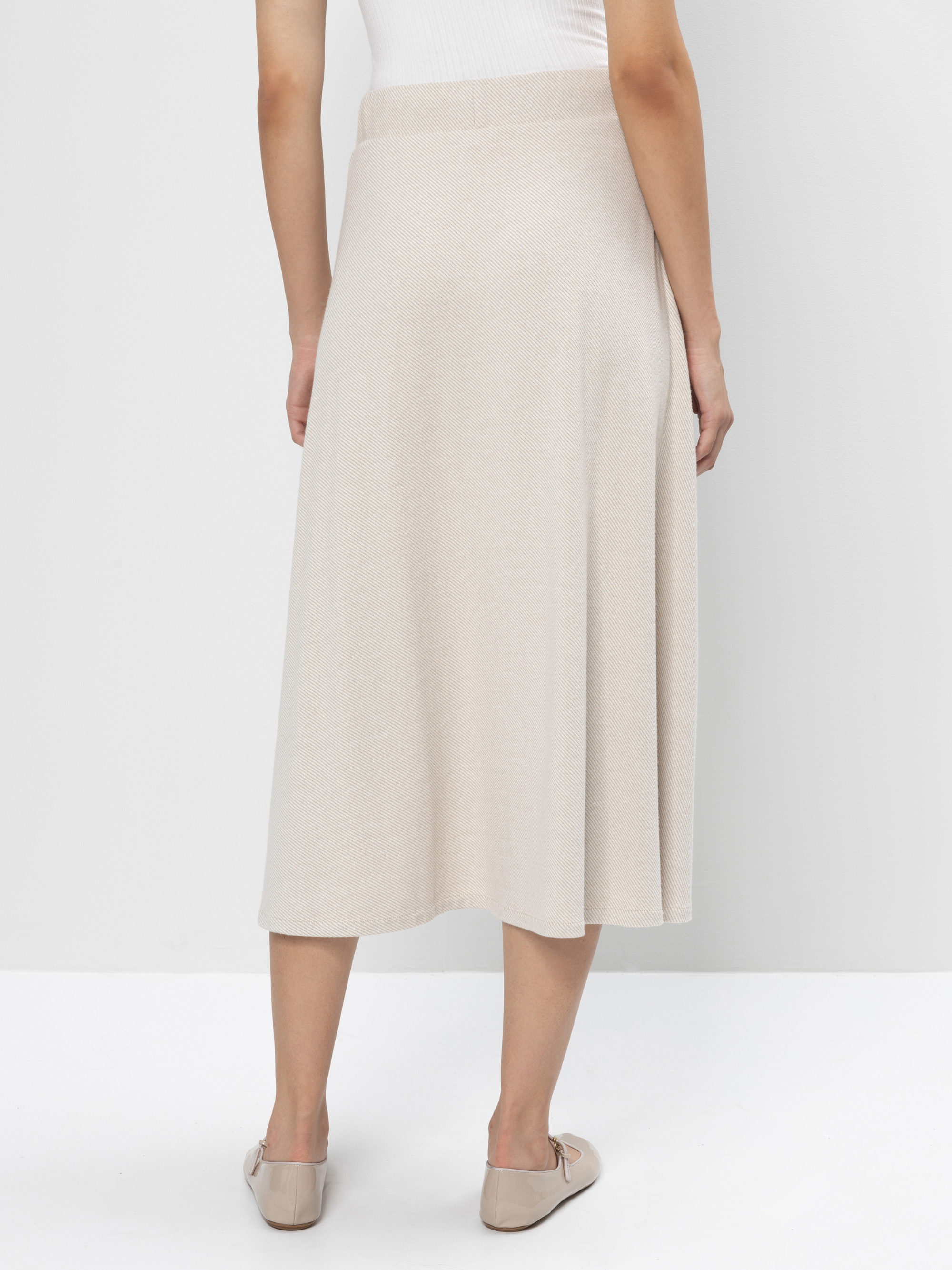 Eine Person steht mit dem Rücken zur Kamera, trägt einen hellbeigen, knielangen Rock in A-Linie, ein weißes ärmelloses Oberteil und hellbeige flache Schuhe. Der Hintergrund ist schlicht und weiß.