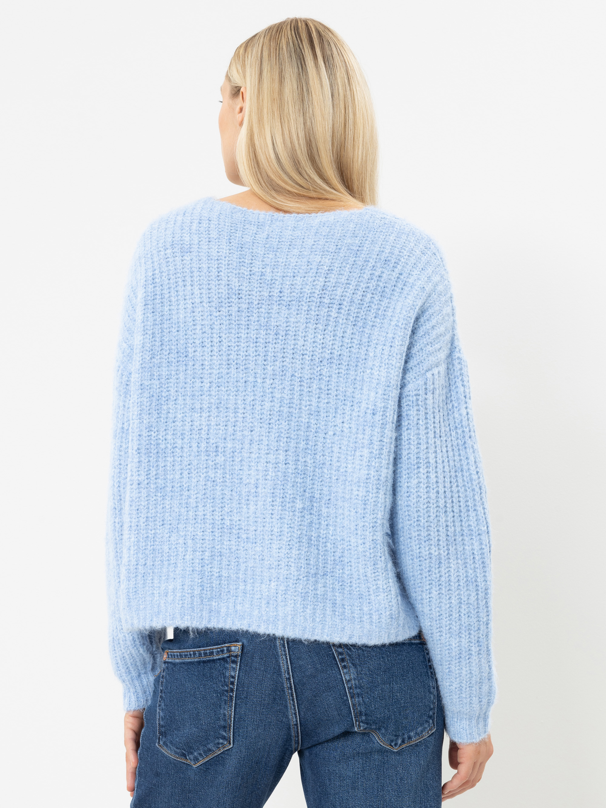 Eine Frau mit langen blonden Haaren, von hinten gesehen, trägt einen hellblauen Strickpullover und eine blaue Jeans vor einem schlichten weißen Hintergrund.