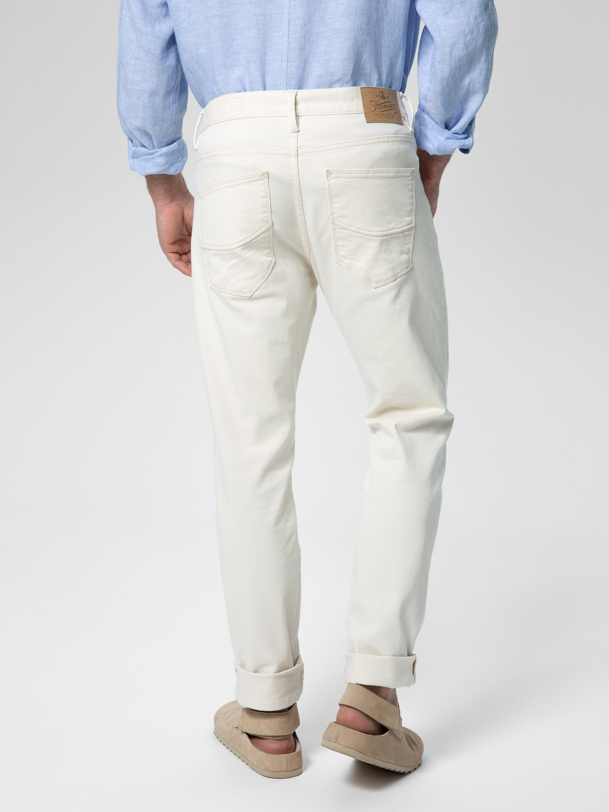 Eine Person, die ein hellblaues Hemd und eine hochgekrempelte cremefarbene Jeans trägt, steht mit dem Gesicht zur Seite und zeigt die Gesäßtaschen und einen braunen Lederaufnäher am Bund. Die Person trägt außerdem beigefarbene Sandalen.