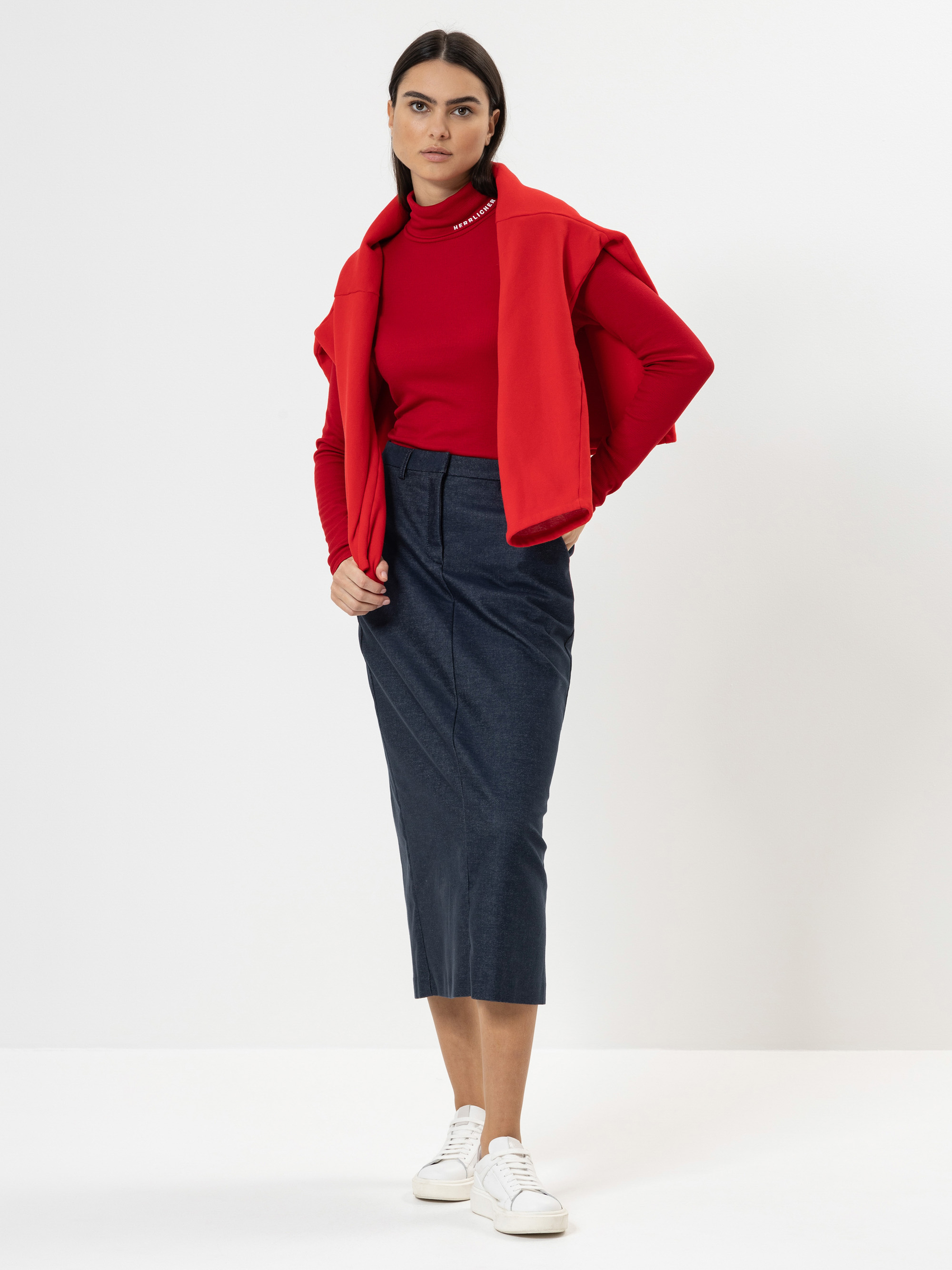 Eine Frau steht vor einem schlichten weißen Hintergrund und trägt ein rotes langärmeliges Oberteil, einen roten Pullover, der ihr über die Schultern fällt, einen dunkelblauen Midirock und weiße Turnschuhe. Sie hat ihr dunkles Haar zurückgebunden un