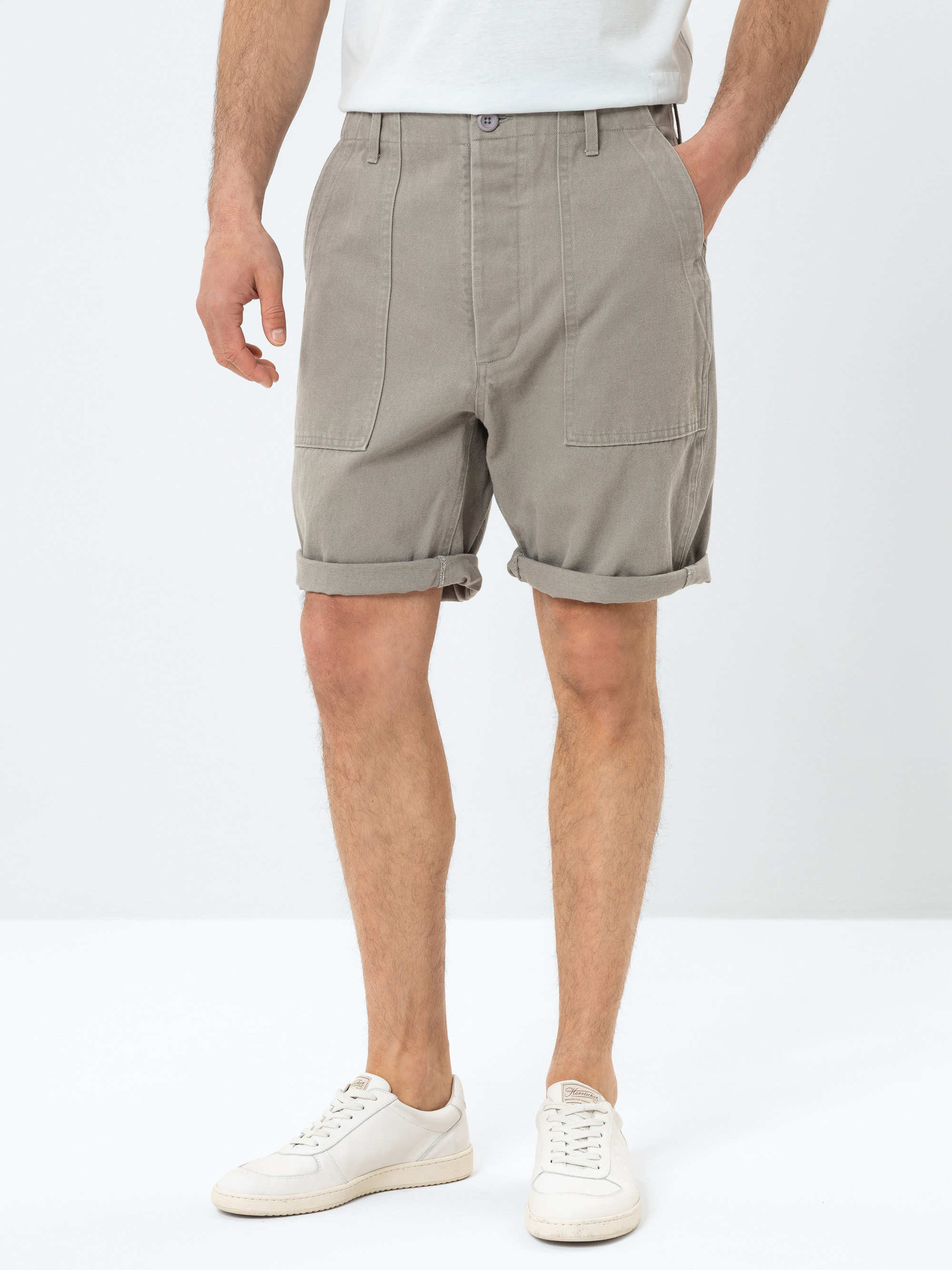 Eine Person, die ein weißes T-Shirt, aufgerollte graue Shorts mit Vordertaschen und weiße Turnschuhe trägt, steht vor einem schlichten weißen Hintergrund. Das Bild ist an den Schultern abgeschnitten und konzentriert sich auf das Outfit.