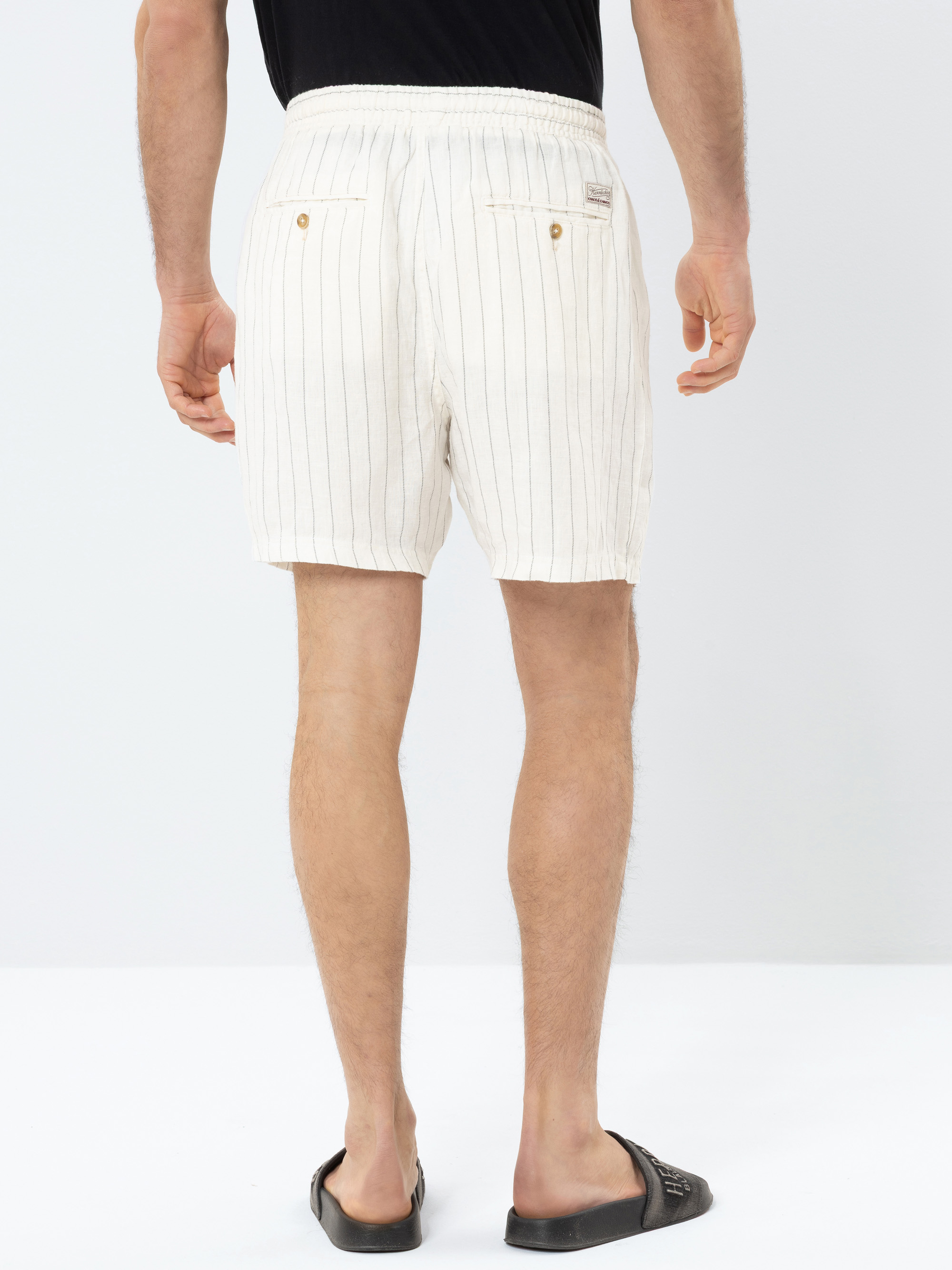 Eine Person, die weiße Shorts mit dünnen vertikalen Streifen, ein schwarzes Hemd und schwarze Gleitsandalen trägt, steht mit dem Gesicht zur Kamera vor einem schlichten weißen Hintergrund.