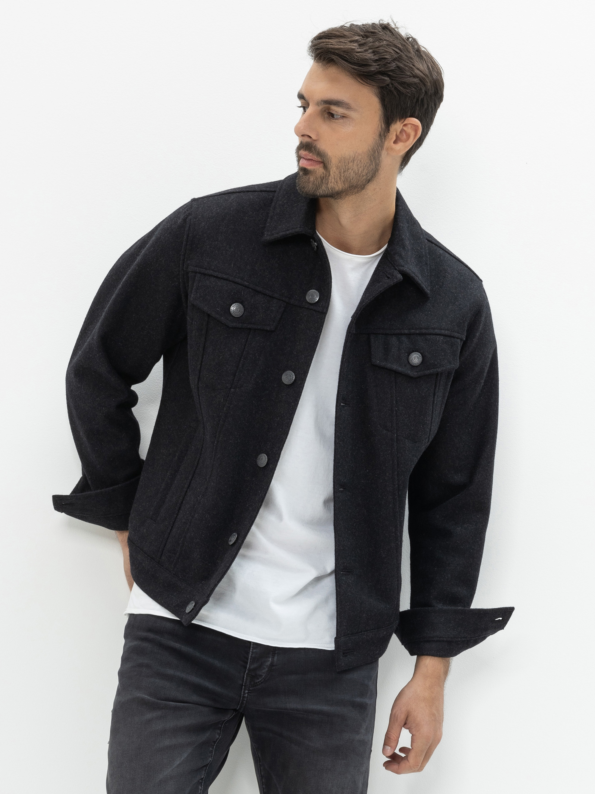Ein Mann mit kurzen dunklen Haaren und gestutztem Bart trägt eine schwarze Jacke mit Knöpfen über einem weißen T-Shirt und schwarze Jeans. Er steht vor einem schlichten weißen Hintergrund und schaut nach links.