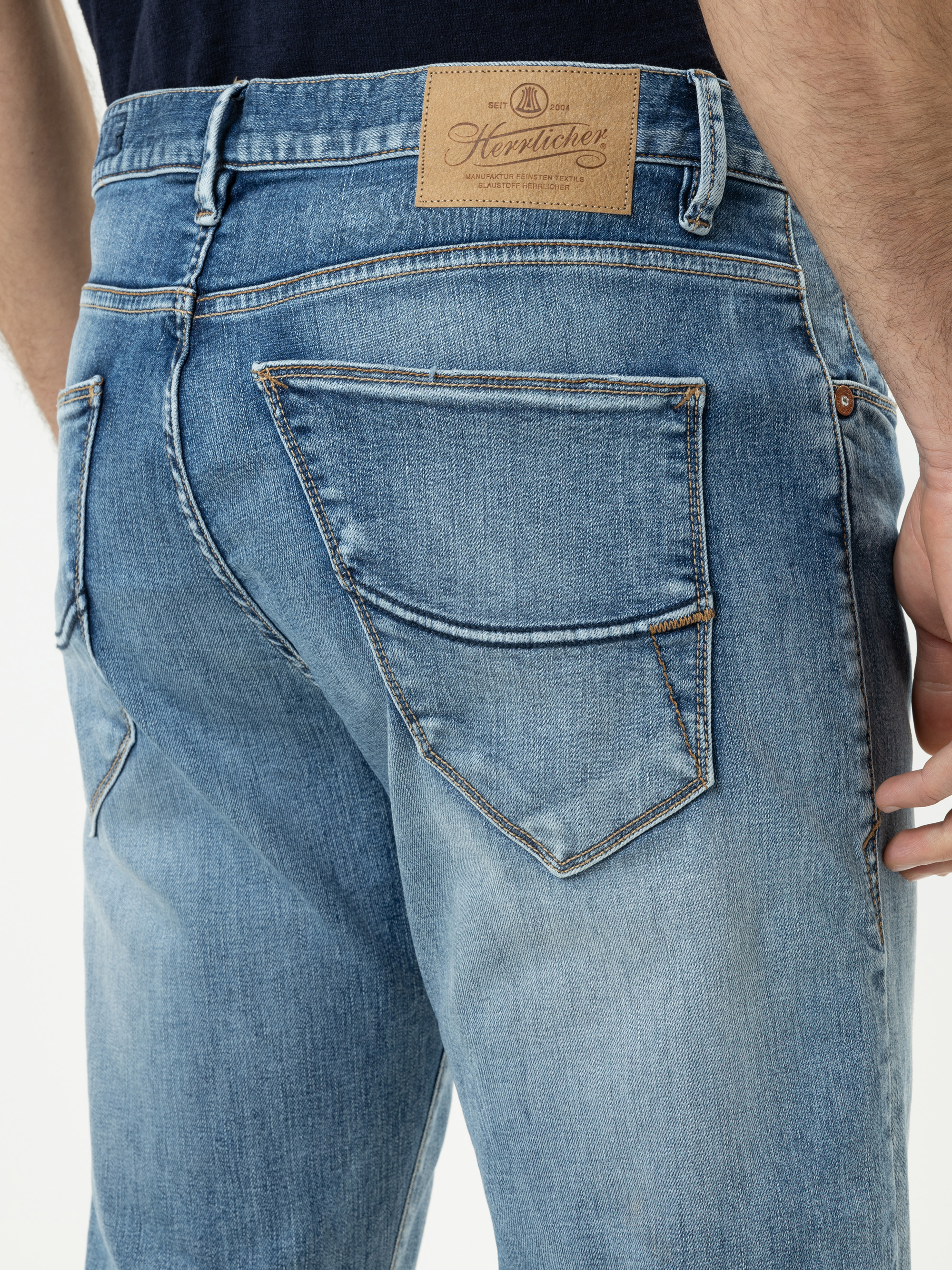 Nahaufnahme einer Person, die eine hellblaue Denim-Jeans trägt, mit Gesäßtasche, Nähten und einem hellbraunen Lederaufnäher am Bund mit dem Markennamen "Heritage". Der Hintergrund ist weiß.