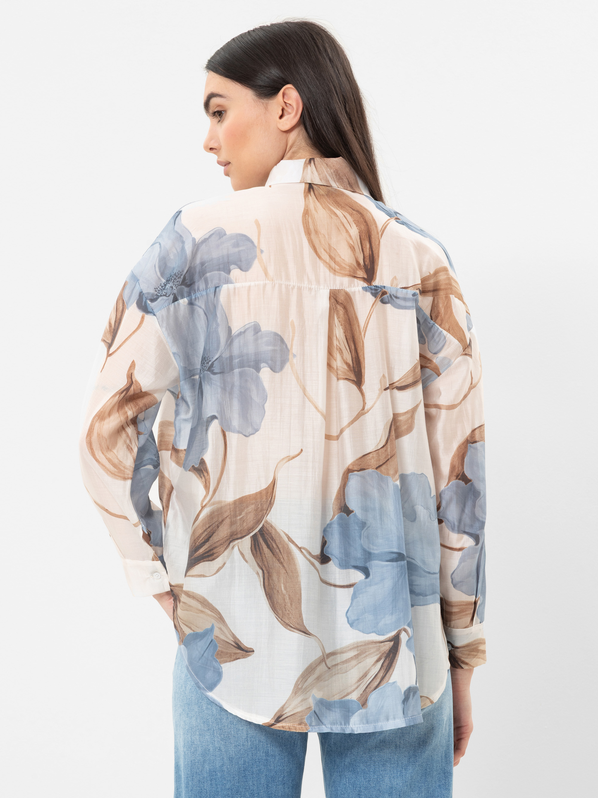 Eine Frau mit langen, dunklen Haaren blickt nach vorne und trägt eine cremefarbene Bluse mit einem großen blauen und braunen Blumenmuster. Außerdem trägt sie eine hellblaue Jeans vor einem schlichten weißen Hintergrund.