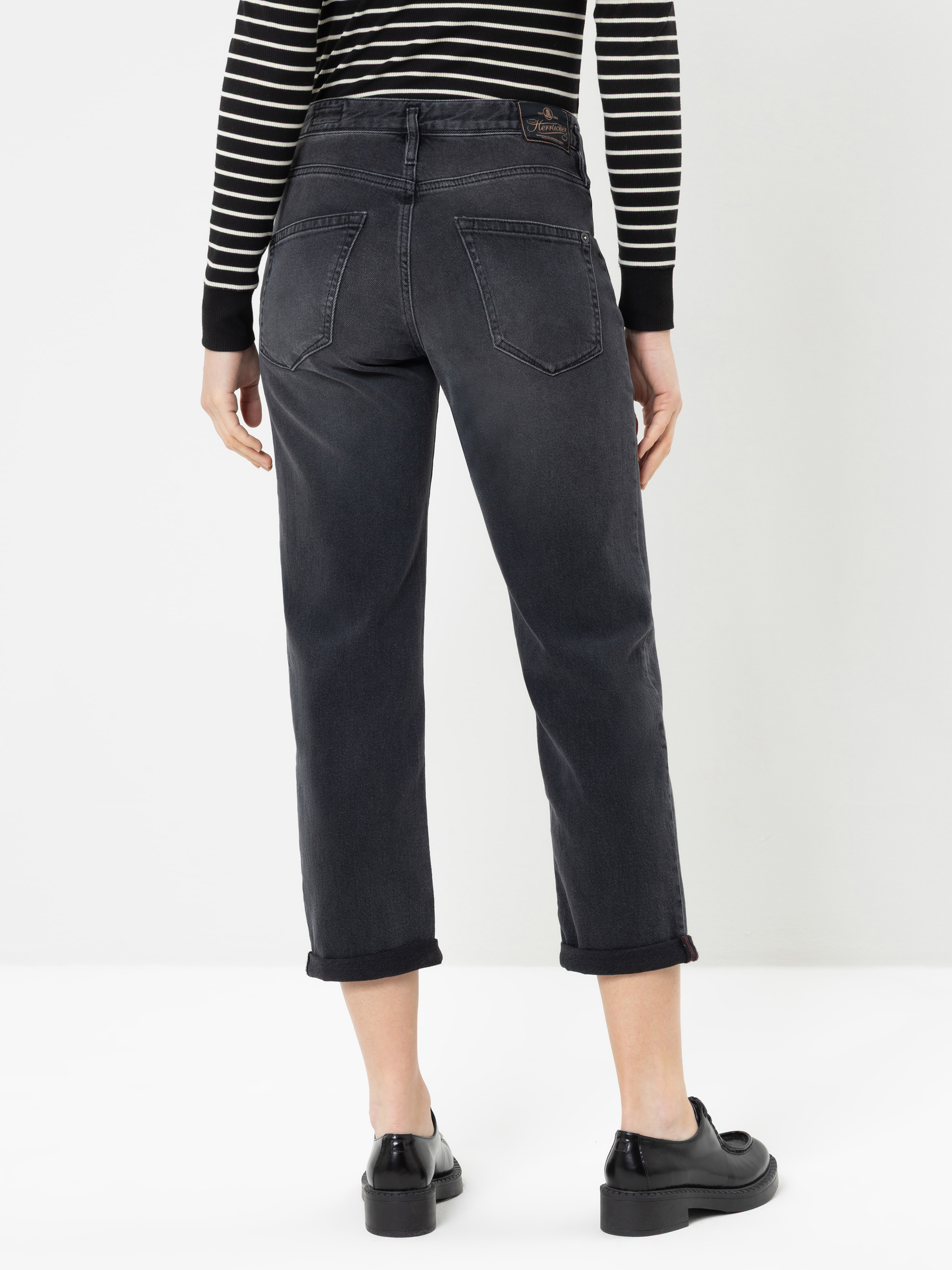 Eine Person, die schwarze Jeans mit Umschlag, schwarze Slipper und ein schwarz-weiß gestreiftes Langarmhemd trägt und mit dem Rücken zur Kamera vor einem schlichten weißen Hintergrund steht.