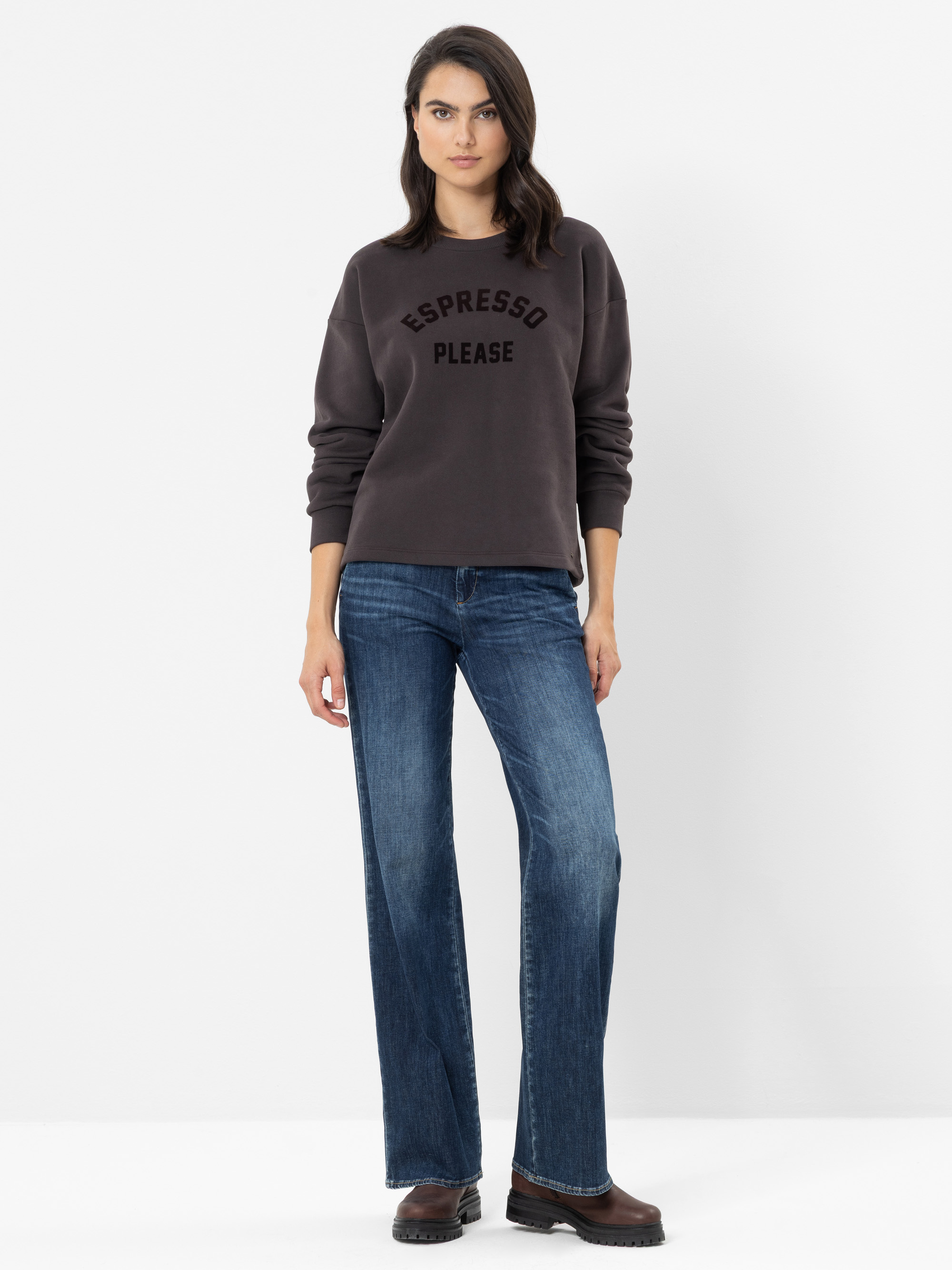 Eine Frau steht vor einem weißen Hintergrund und trägt ein dunkles Sweatshirt mit der Aufschrift „ESPRESSO PLEASE“, eine blaue Jeans mit weitem Bein und braune Schuhe. Sie hat glattes, dunkles Haar und einen entspannten Gesichtsausdruck, wobei ihre