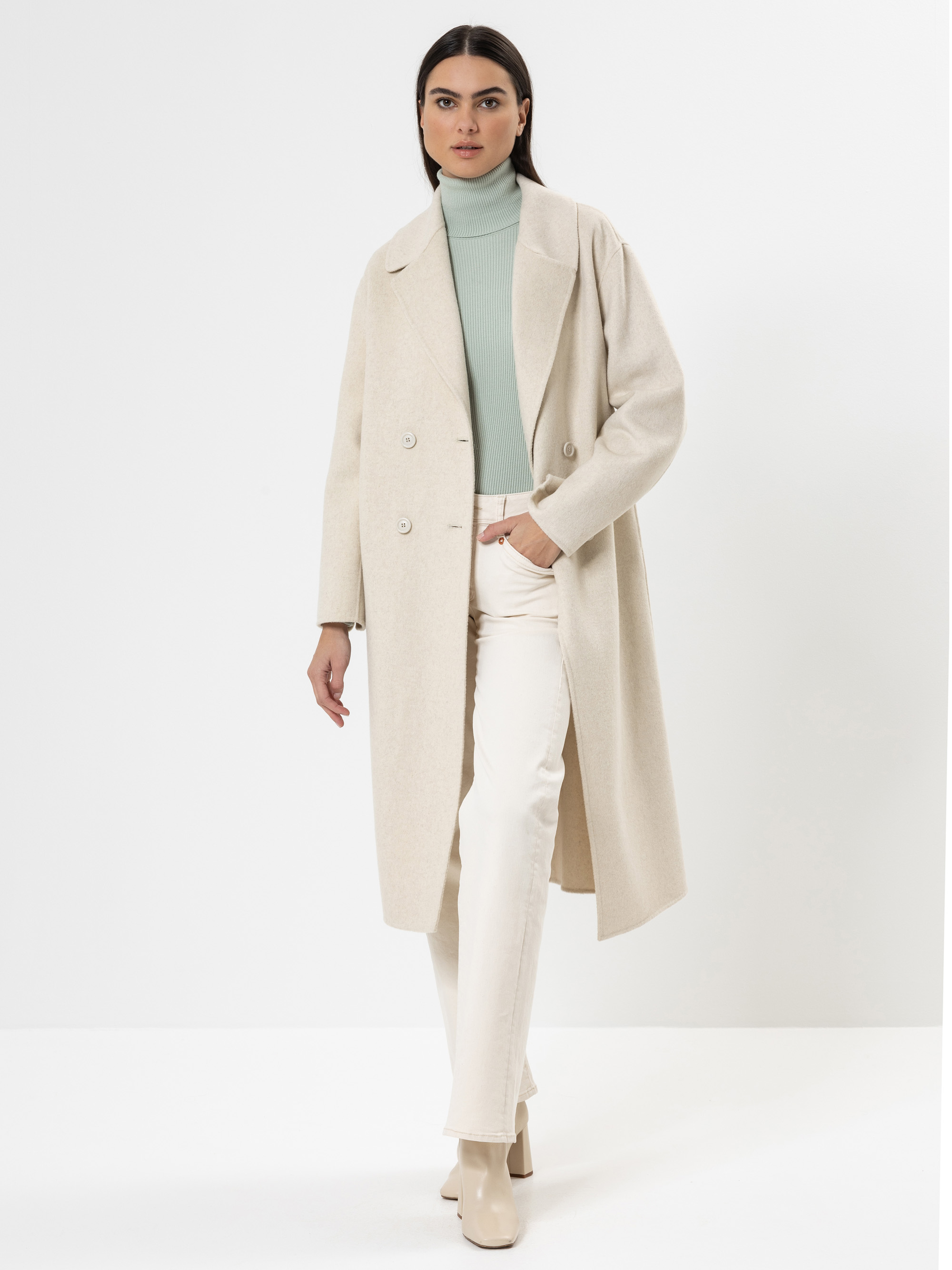 Eine Frau steht vor einem weißen Hintergrund und trägt einen langen, hellbeigen Mantel über einem mintgrünen Rollkragenpullover, eine cremefarbene Hose und beigefarbene Stiefeletten, eine Hand in der Tasche.