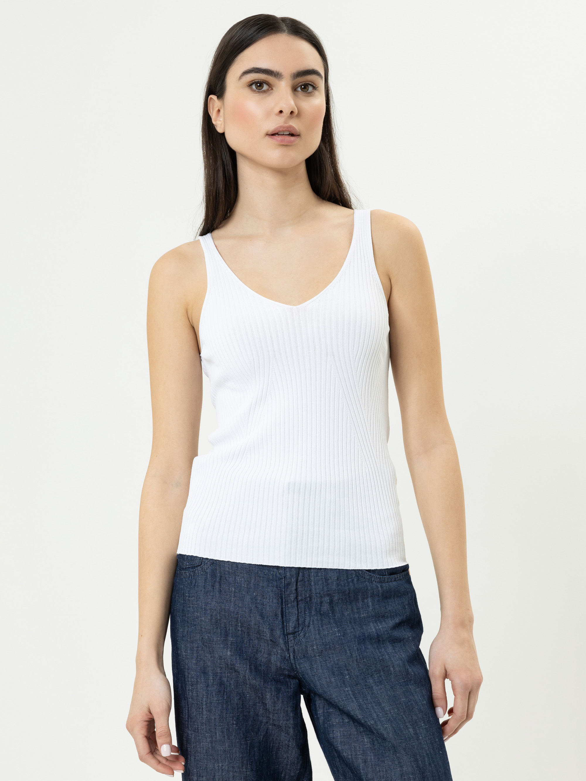 Eine Frau mit langen dunklen Haaren trägt ein weißes ärmelloses geripptes Tank-Top und dunkelblaue Jeans. Sie steht vor einem schlichten hellen Hintergrund und blickt leicht zur Seite.