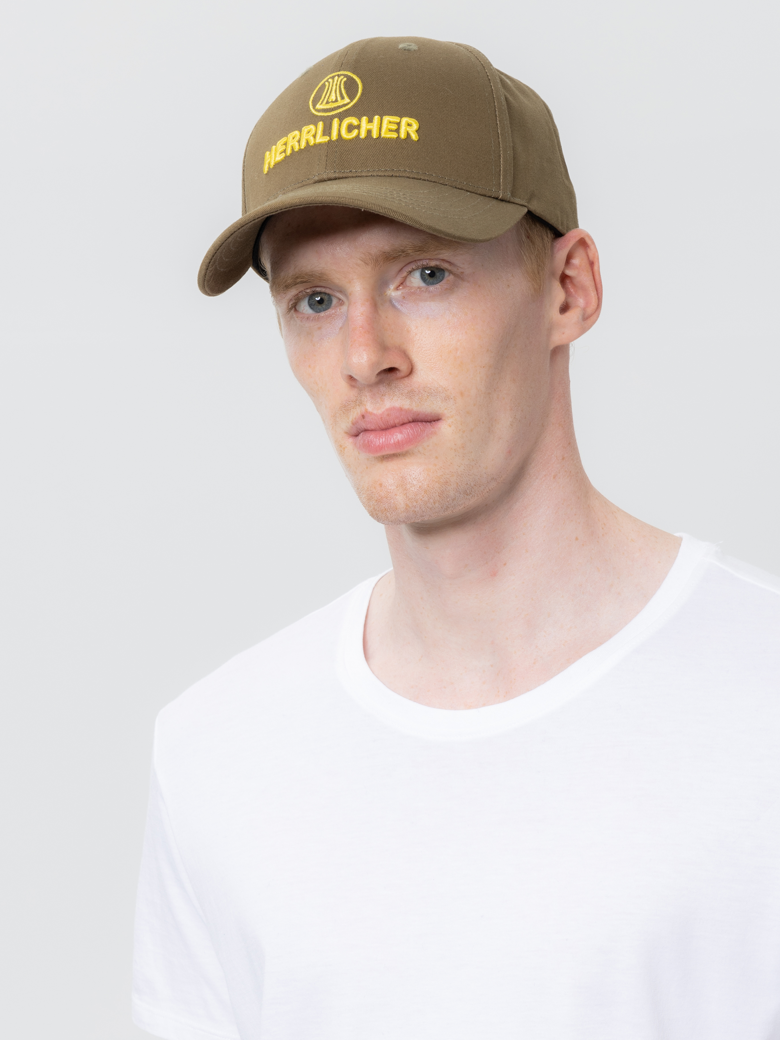 Ein junger Mann mit heller Haut, der eine khakifarbene Baseballkappe mit gelber Aufschrift HERRENLICHER und ein schlichtes weißes T-Shirt trägt, posiert vor einem hellgrauen Hintergrund.