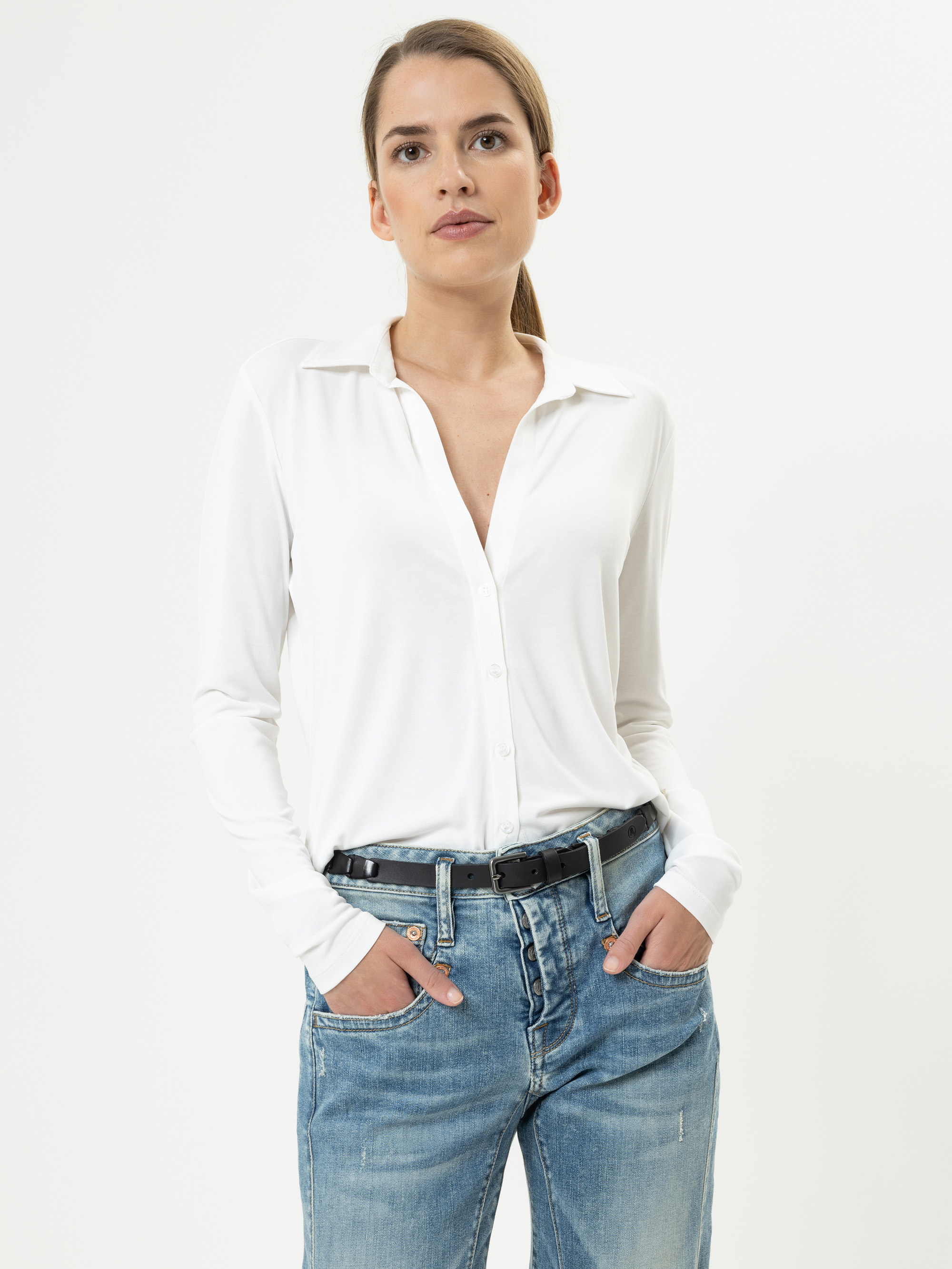 Eine Frau posiert vor einem schlichten weißen Hintergrund. Sie trägt ein weißes Button-up-Hemd, dessen oberste Knöpfe offen sind, hellblaue Jeans und einen schwarzen Gürtel. Ihre Hände stecken in den Taschen und sie hat einen neutralen Gesichtsausdr