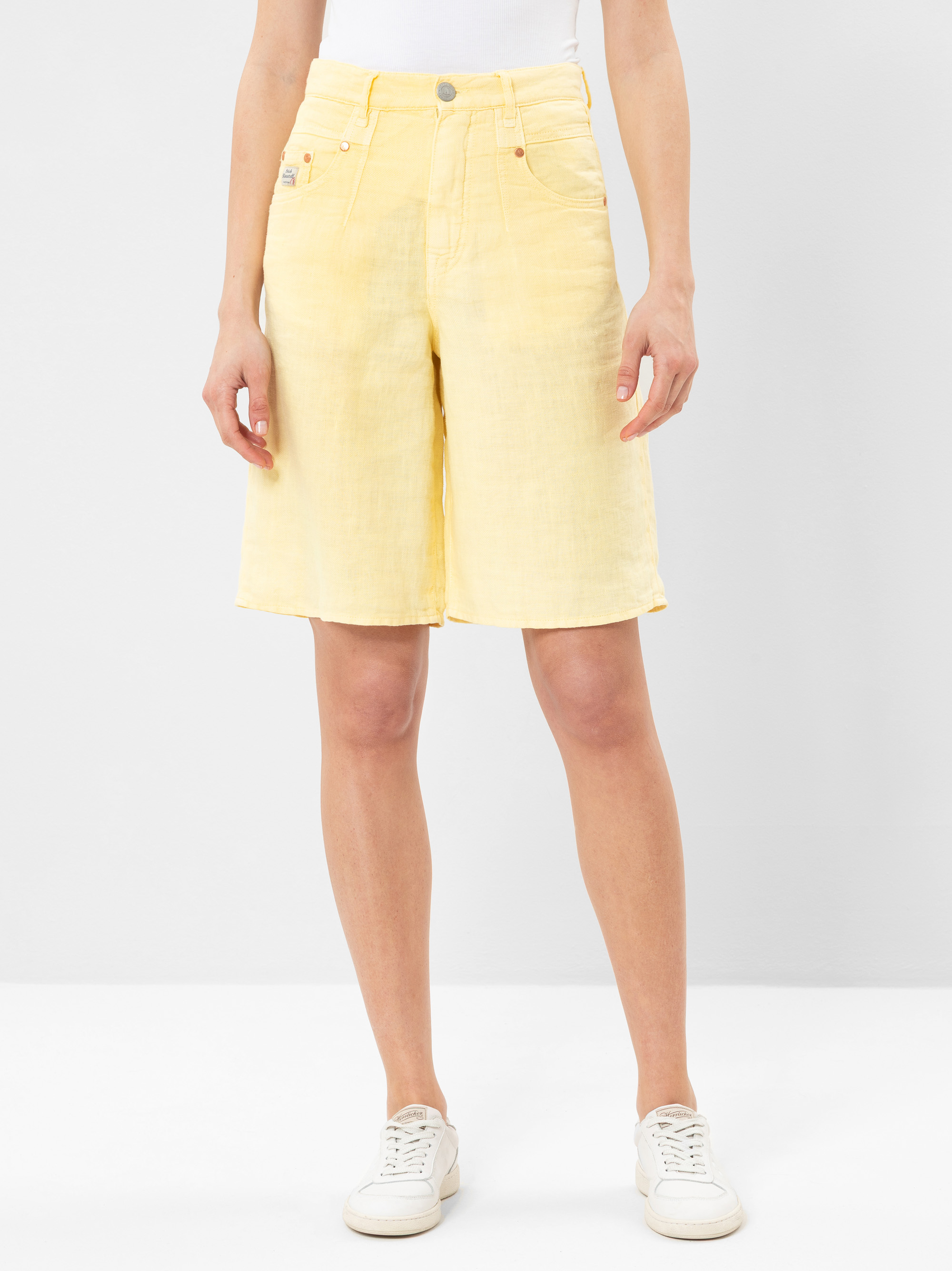Eine Person, die blassgelbe knielange Shorts, ein weißes Oberteil und weiße Turnschuhe trägt, steht vor einem schlichten hellgrauen Hintergrund. Das Foto zeigt ihre Beine und ihren Oberkörper, die Arme liegen entspannt an ihren Seiten.