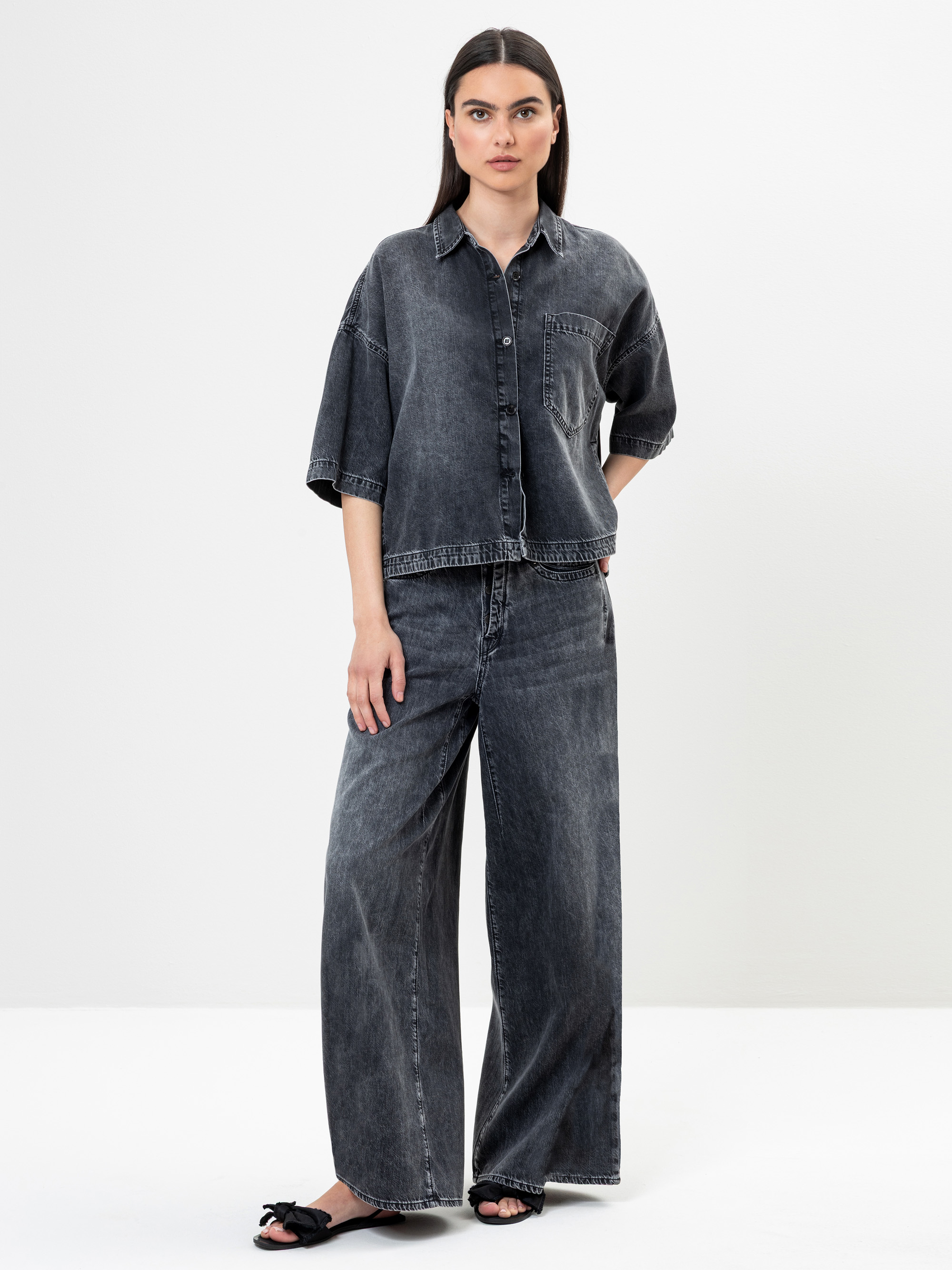 Eine Frau steht vor einem schlichten weißen Hintergrund und trägt ein locker sitzendes, kurzärmeliges, dunkles Jeanshemd und eine dazu passende, weit geschnittene Jeans. Sie hat glattes, dunkles Haar und trägt schwarze flache Sandalen mit Schleifen.