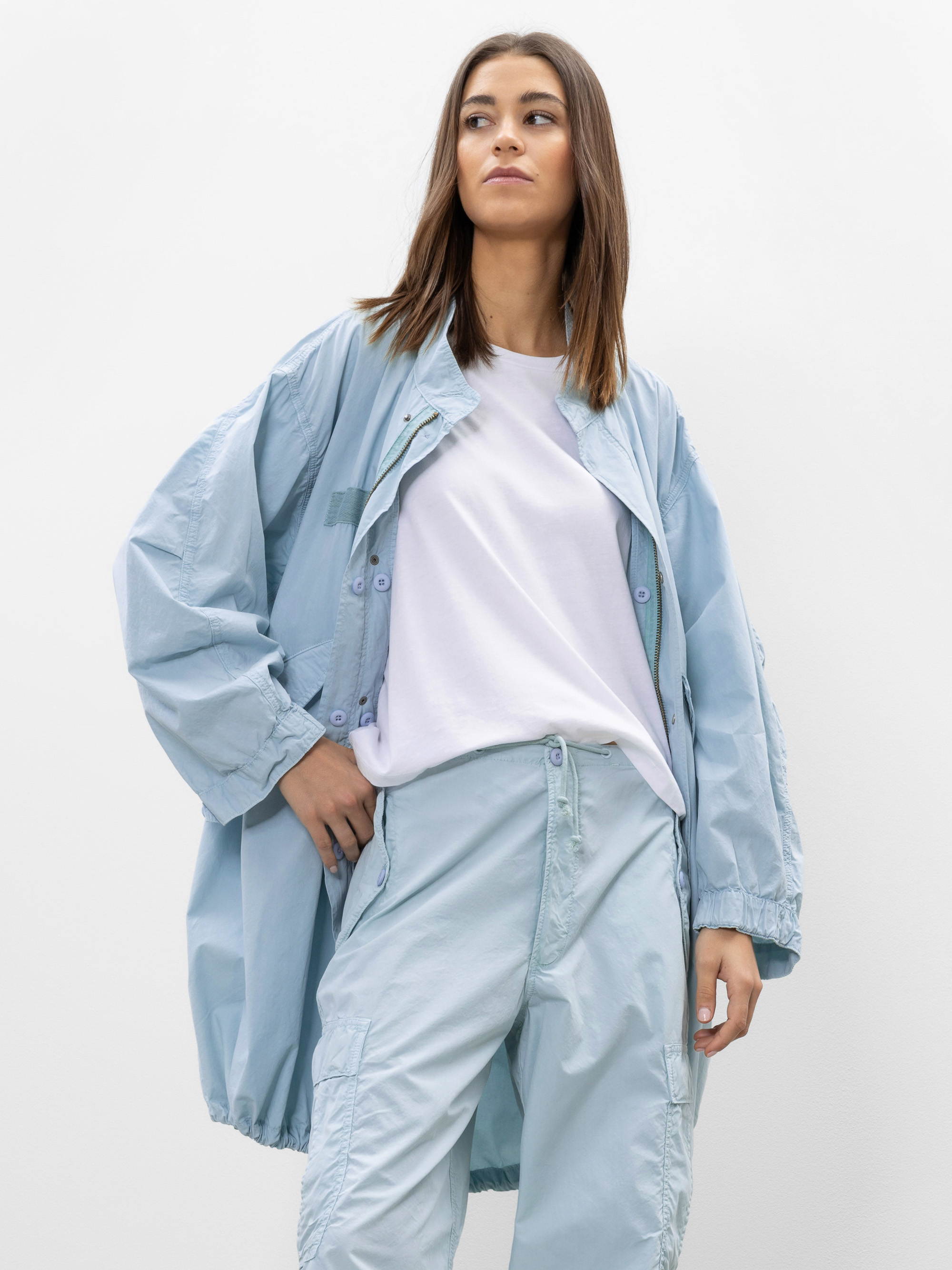 Eine Frau mit glattem braunem Haar trägt eine hellblaue übergroße Jacke, eine dazu passende hellblaue Hose und ein weißes T-Shirt und steht vor einem schlichten weißen Hintergrund.