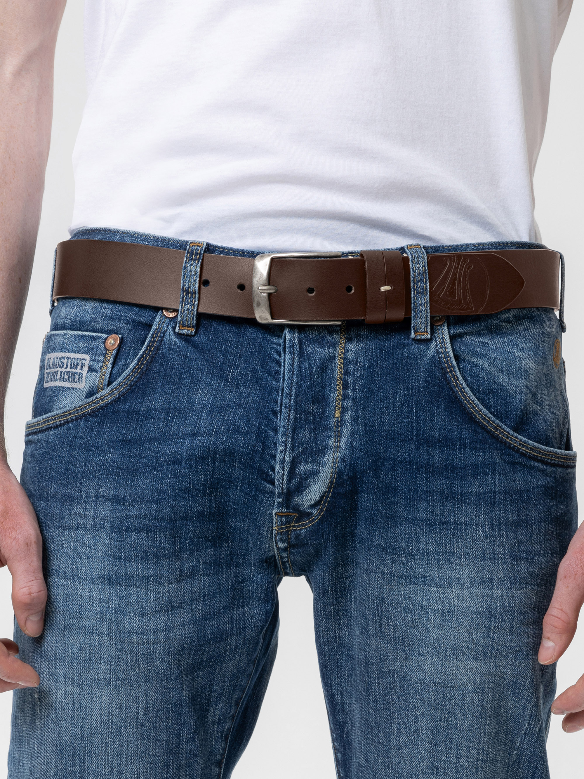 Eine Person, die ein weißes T-Shirt, eine blaue Jeans und einen braunen Ledergürtel mit einer silbernen Schnalle trägt, wird von der Taille bis zur Mitte des Oberschenkels gezeigt, wobei die Hände in der Nähe der Taschen ruhen.