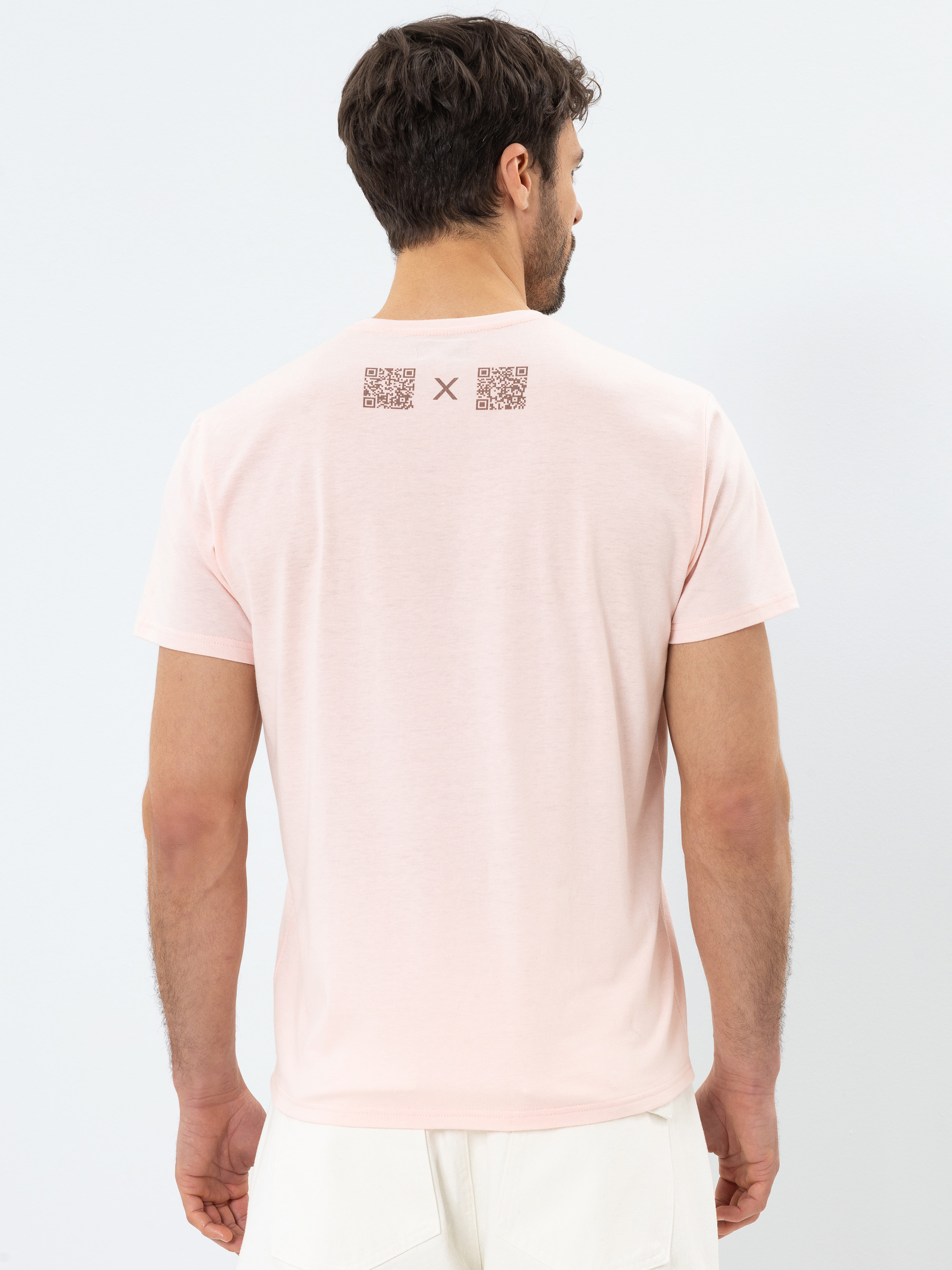 Ein Mann, der ein hellrosa T-Shirt und weiße Shorts trägt, wird von hinten gezeigt. Auf dem T-Shirt sind zwei QR-Codes und ein kleines X in der Nähe des Halsausschnitts aufgedruckt. Der Hintergrund ist einfarbig und hell gefärbt.