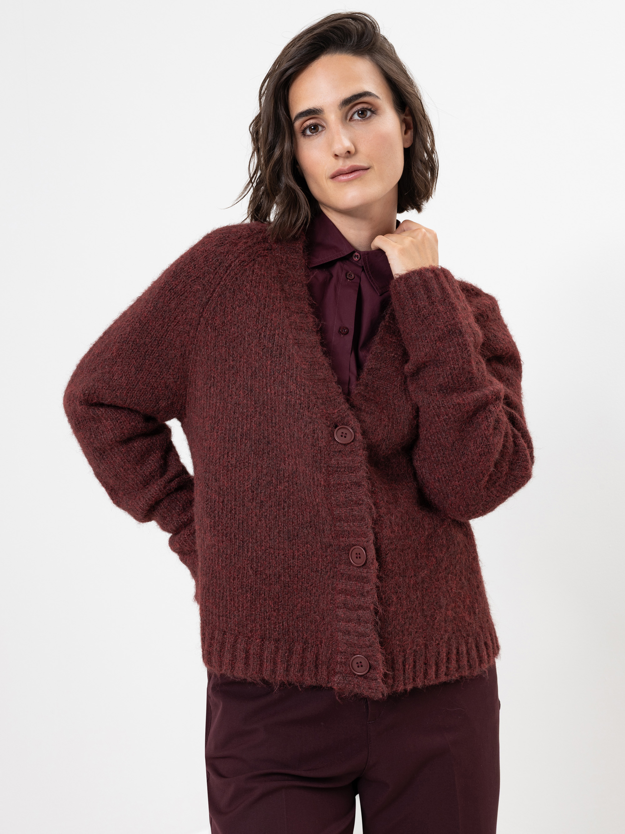 Eine Frau mit kurzen braunen Haaren trägt eine kastanienbraune, strukturierte Strickjacke mit Knopfleiste über einem passenden kastanienbraunen Hemd mit Kragen und einer Hose und steht vor einem schlichten weißen Hintergrund.