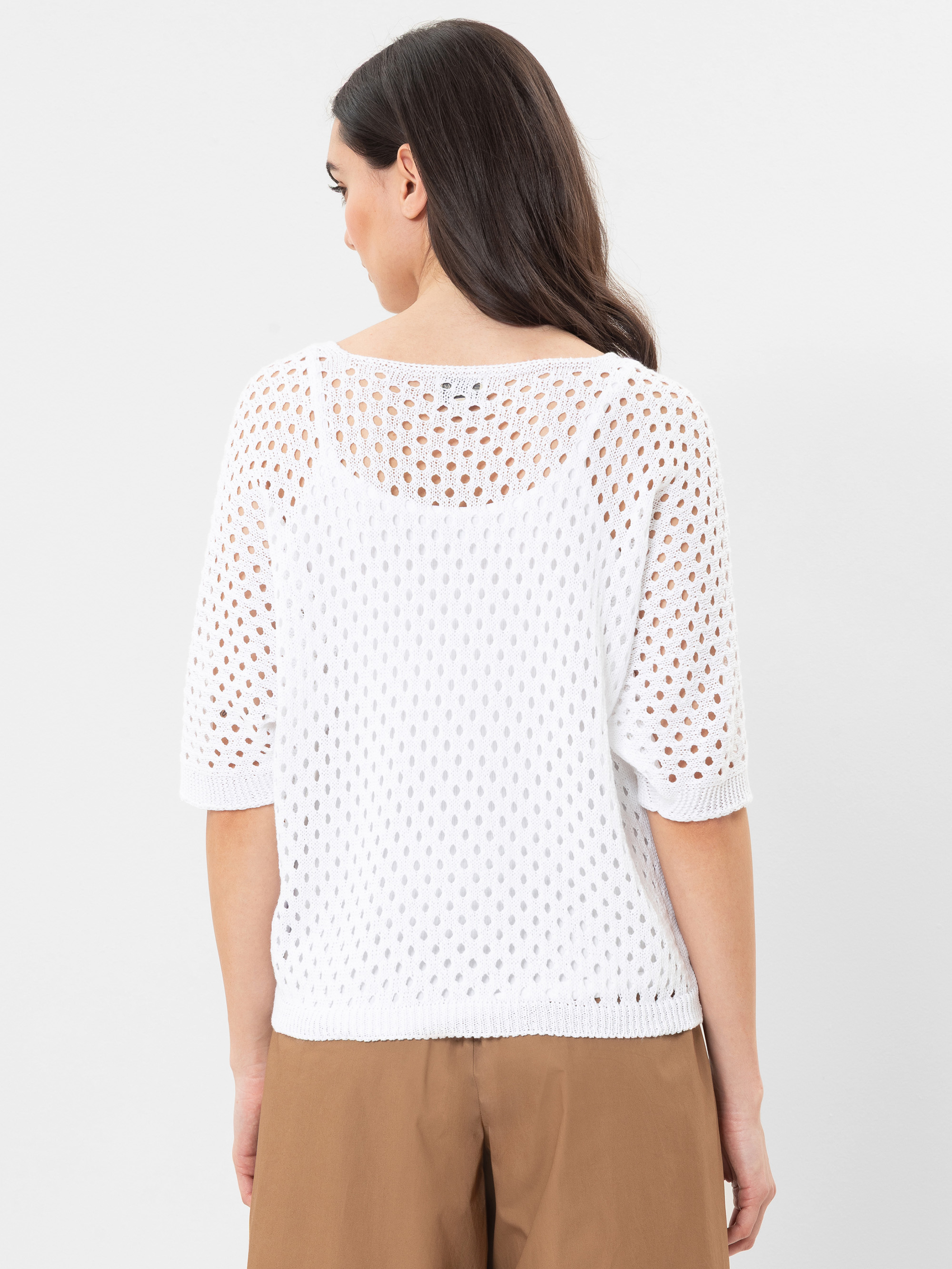 Eine Frau mit langen dunklen Haaren wird von hinten gezeigt. Sie trägt ein weißes, kurzärmeliges, gehäkeltes Strickoberteil über einem weißen Unterhemd und eine hellbraune Hose. Der Hintergrund ist einfarbig weiß.