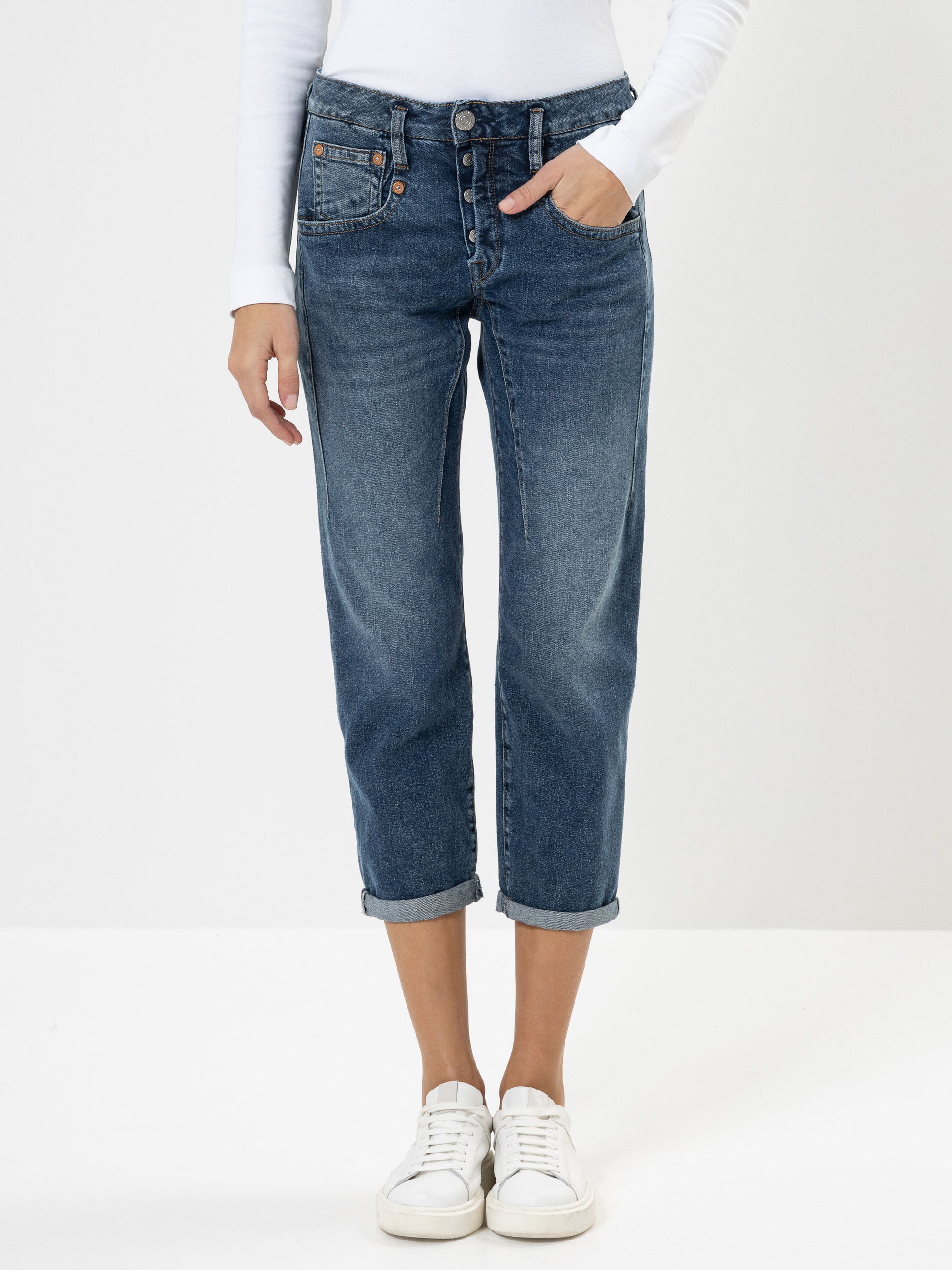 Eine Person, die eine blaue Jeans mit Manschetten, ein langärmeliges weißes Hemd und weiße Turnschuhe trägt, steht vor einem schlichten weißen Hintergrund und hat eine Hand in der Tasche.