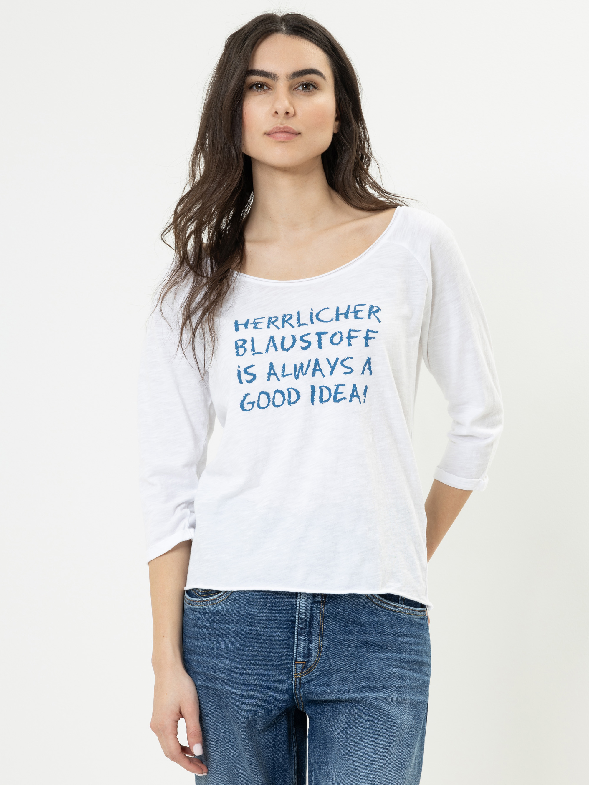 Frau mit langen dunklen Haaren, die ein weißes Hemd mit blauer Aufschrift "Herrlicher Blaustoff ist immer eine gute Idee!" und blaue Jeans trägt und vor einem schlichten weißen Hintergrund steht.