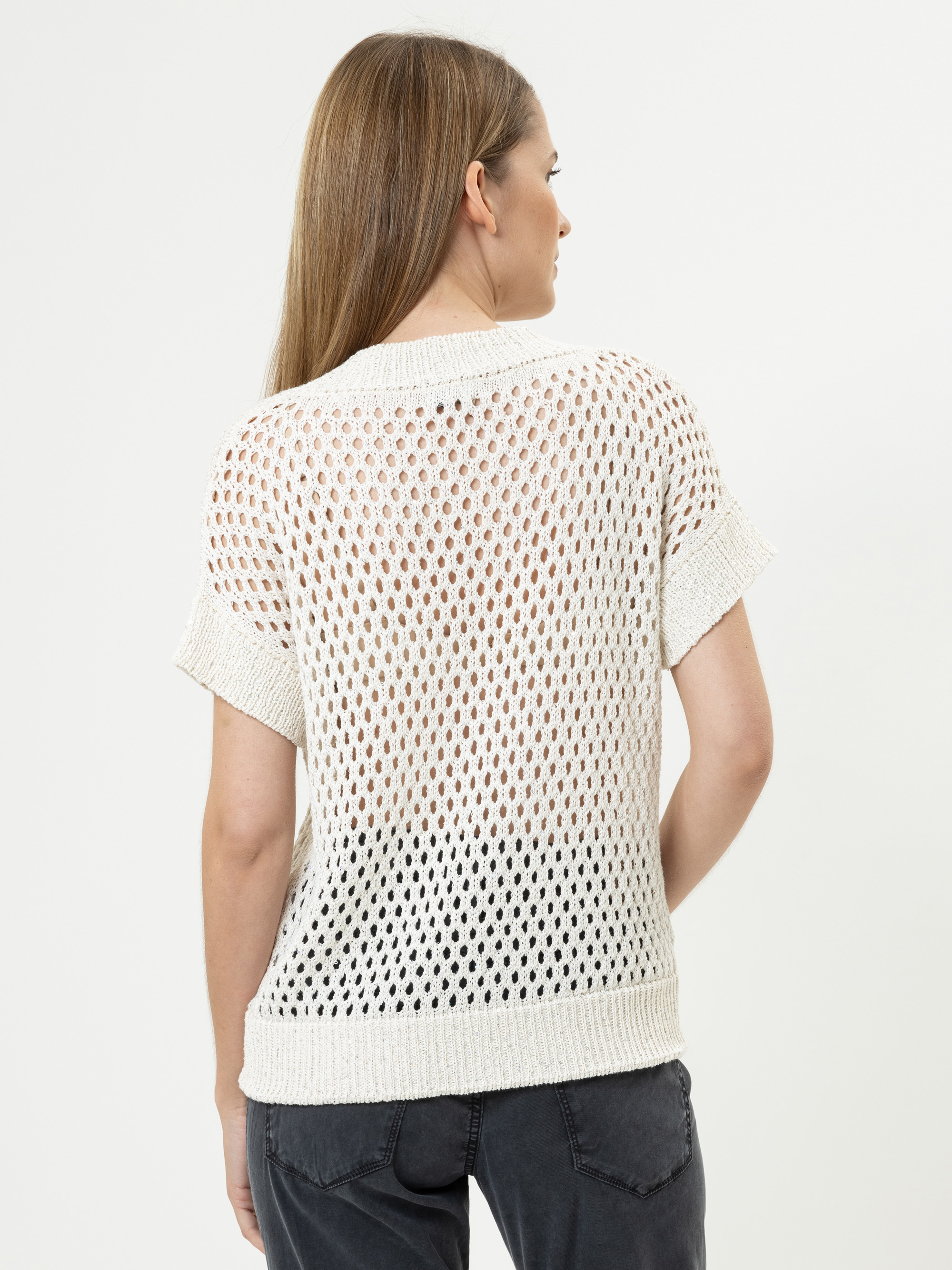 Eine Frau mit glattem, hellbraunem Haar wird von hinten gezeigt. Sie trägt einen kurzärmeligen, weißen, offen gestrickten Pullover über einem weißen Oberteil und eine dunkle Hose und steht vor einem schlichten weißen Hintergrund.