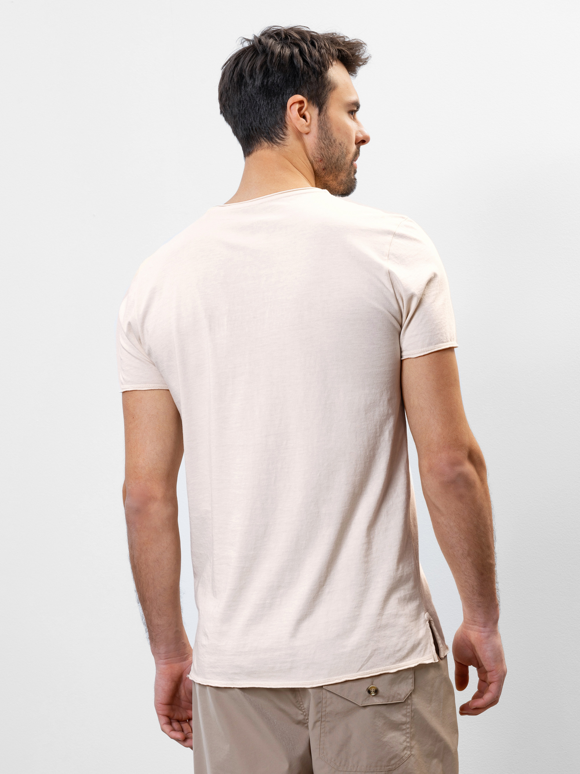 Ein Mann mit kurzen dunklen Haaren steht mit dem Rücken zur Kamera und trägt ein hellbeiges kurzärmeliges T-Shirt und hellbraune Shorts. Der Hintergrund ist einfarbig und hell.