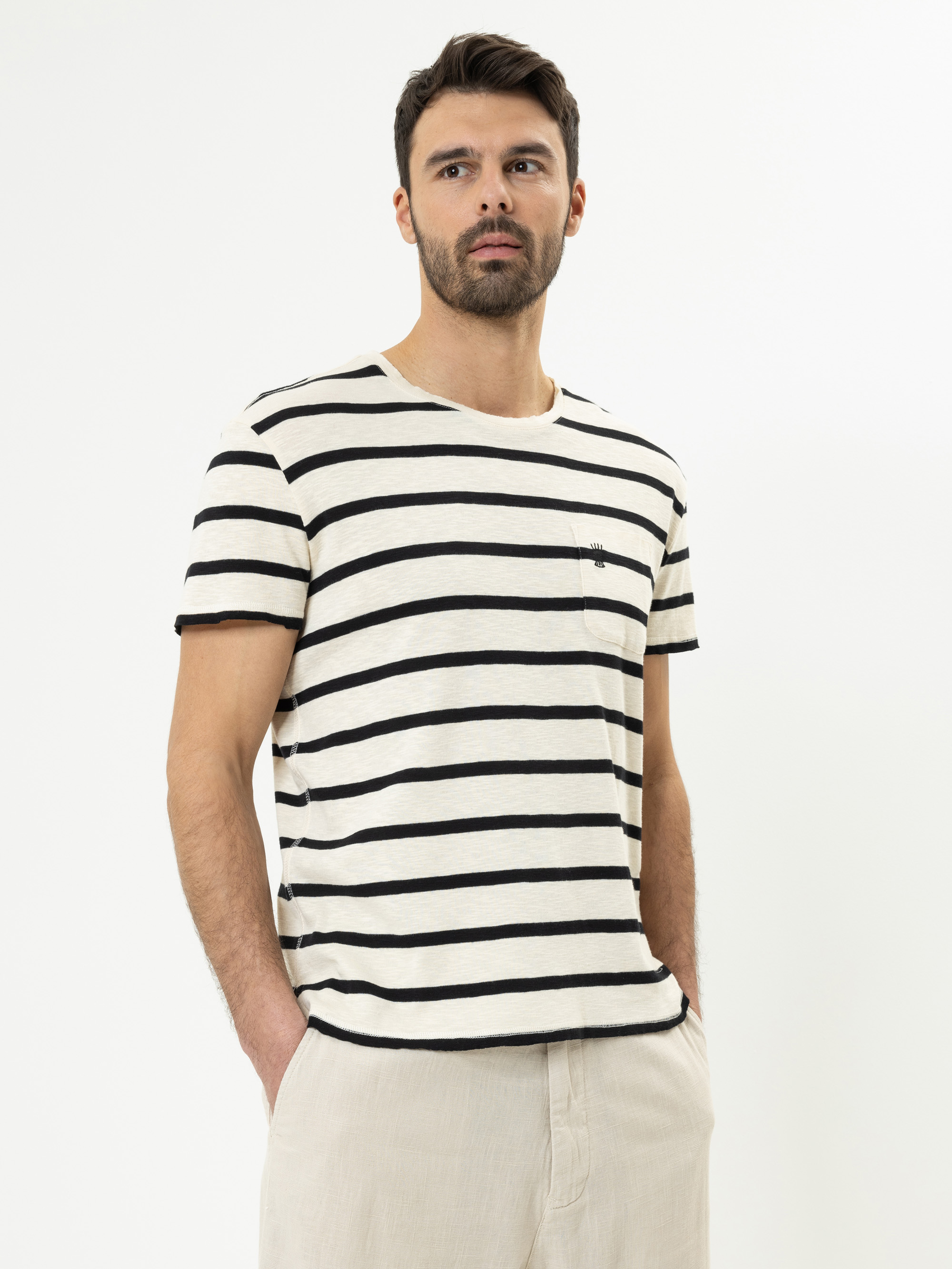 Ein Mann, der ein cremefarbenes T-Shirt mit schwarzen Querstreifen und eine hellbeige Hose trägt, steht vor einem schlichten weißen Hintergrund und schaut leicht zur Seite, die Hände in den Taschen.