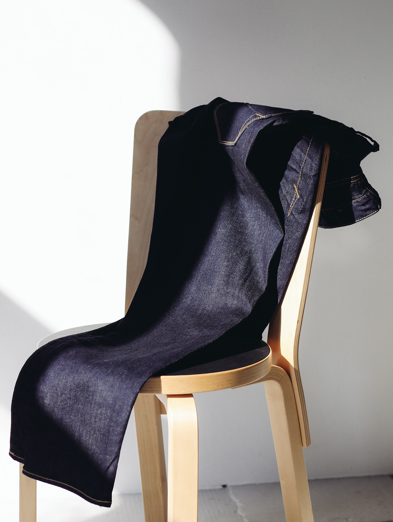 Eine dunkelblaue Jeans, die über die Rückenlehne eines hellen Holzstuhls drapiert war, während das Sonnenlicht weiche Schatten auf die weiße Wand und den Boden warf.