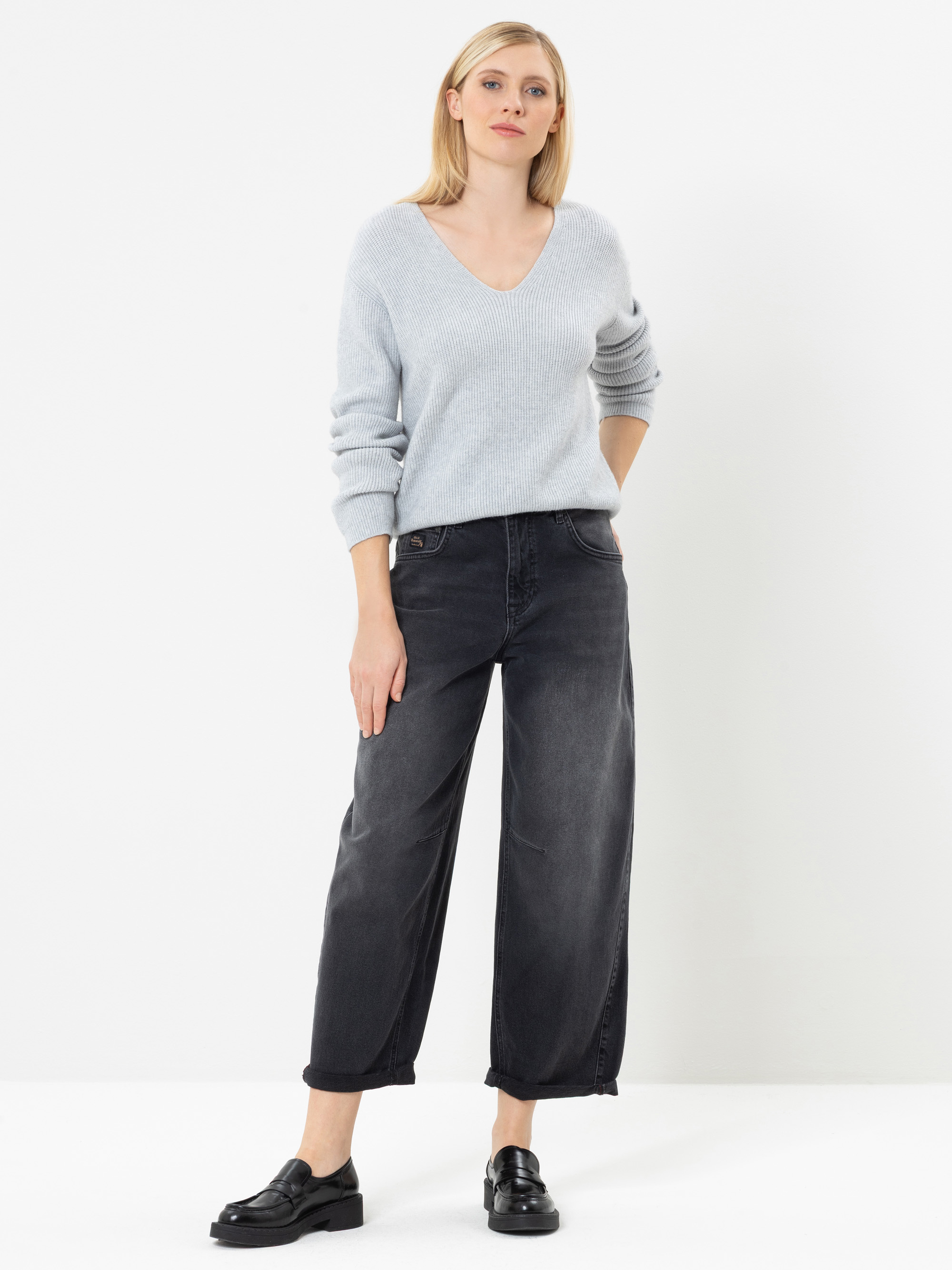 Eine Frau steht vor einem weißen Hintergrund und trägt einen hellgrauen Pullover mit V-Ausschnitt, eine weite schwarze Jeans und schwarze Slipper. Sie hat glattes blondes Haar und steckt ihre linke Hand in die Hosentasche.
