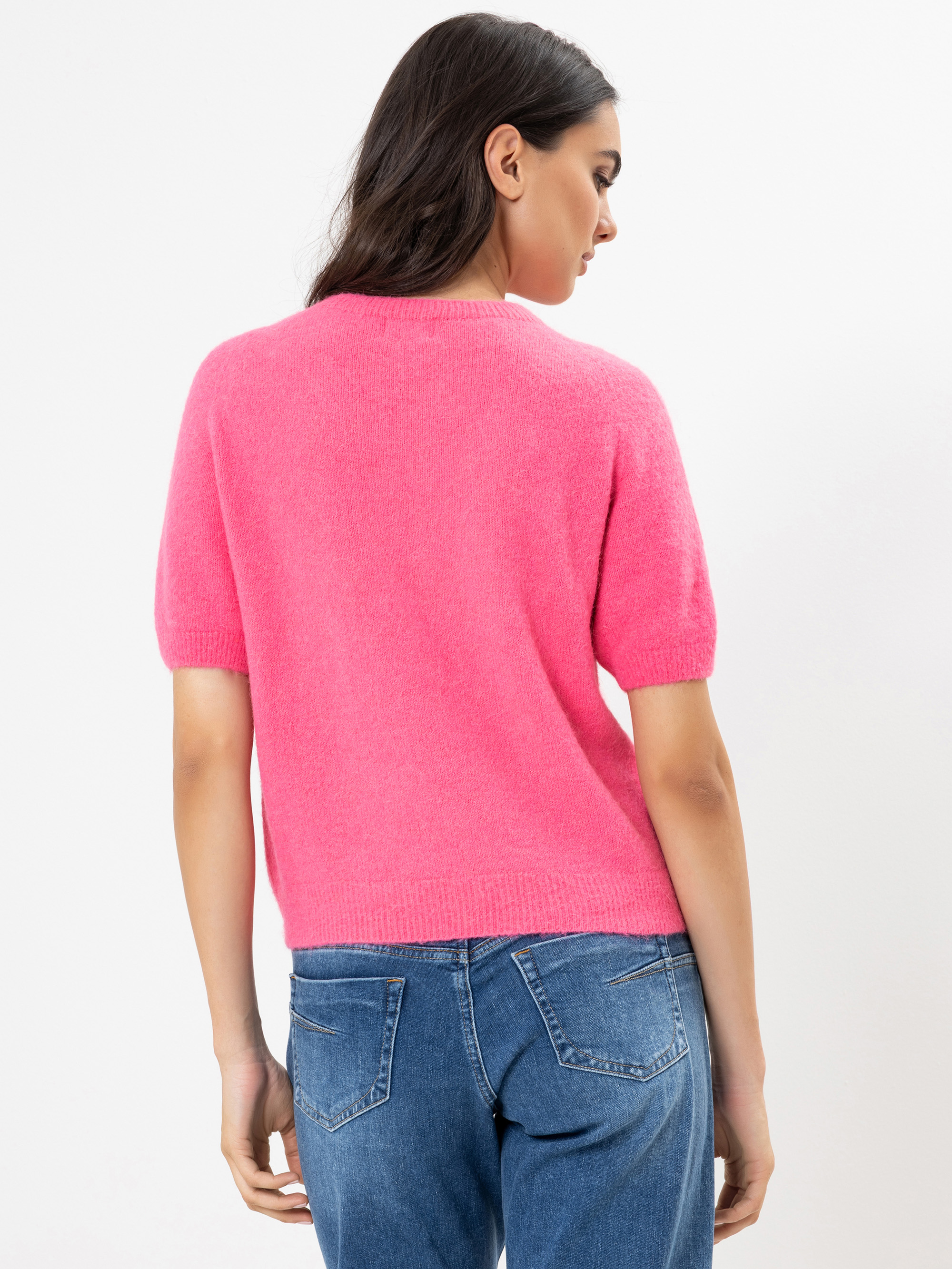 Eine Frau steht mit dem Rücken zur Kamera, trägt einen hellrosa kurzärmeligen Pullover und blaue Jeans vor einem schlichten weißen Hintergrund.