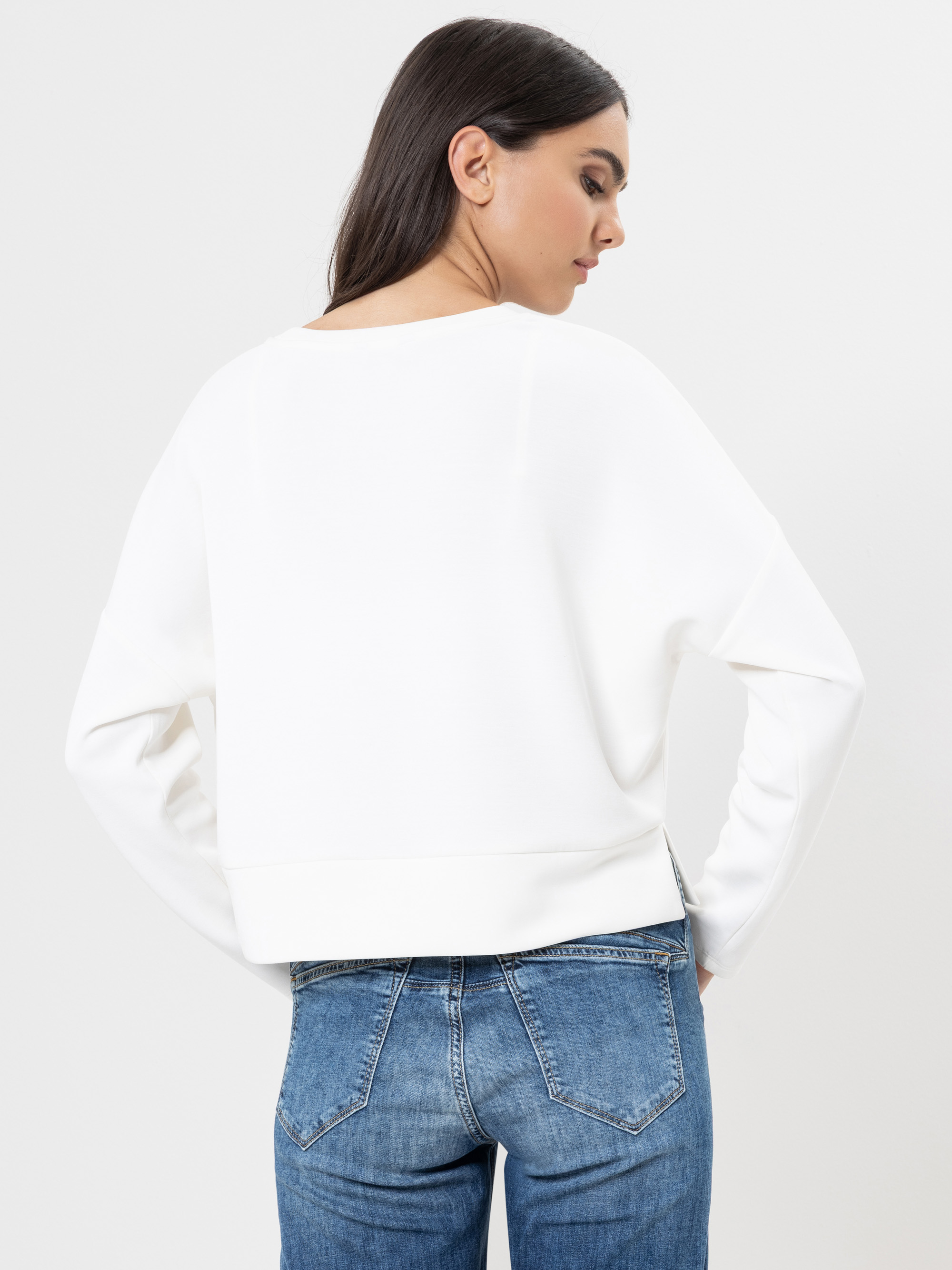 Eine Frau mit langen dunklen Haaren, die ein lockeres weißes Sweatshirt und blaue Jeans trägt, steht mit dem Rücken zur Kamera vor einem schlichten hellen Hintergrund.