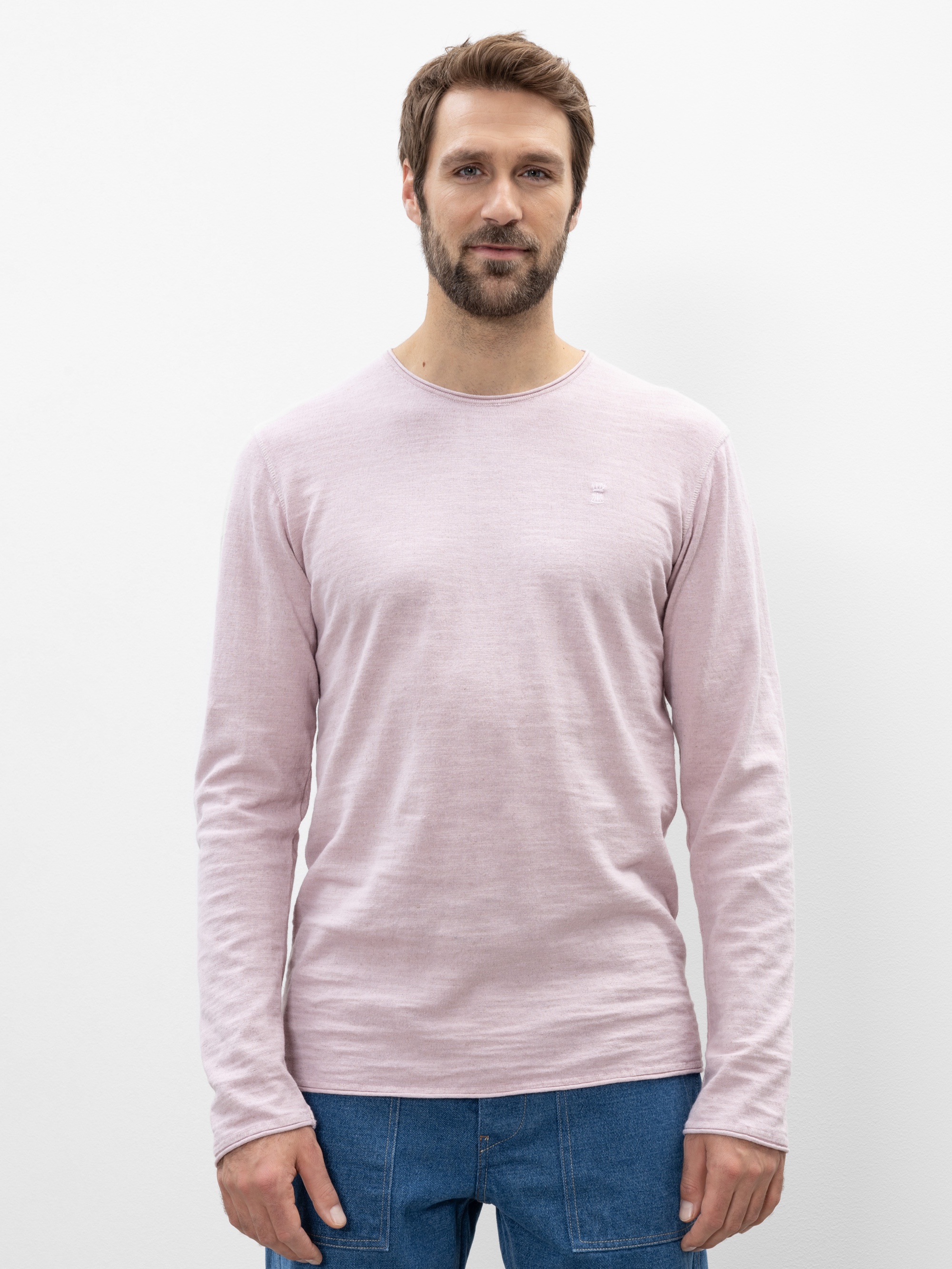 Ein Mann mit kurzen braunen Haaren und einem Bart steht vor einem weißen Hintergrund und trägt ein hellrosa langärmeliges Hemd und blaue Jeans. Er blickt nach vorne und hat einen neutralen Gesichtsausdruck.